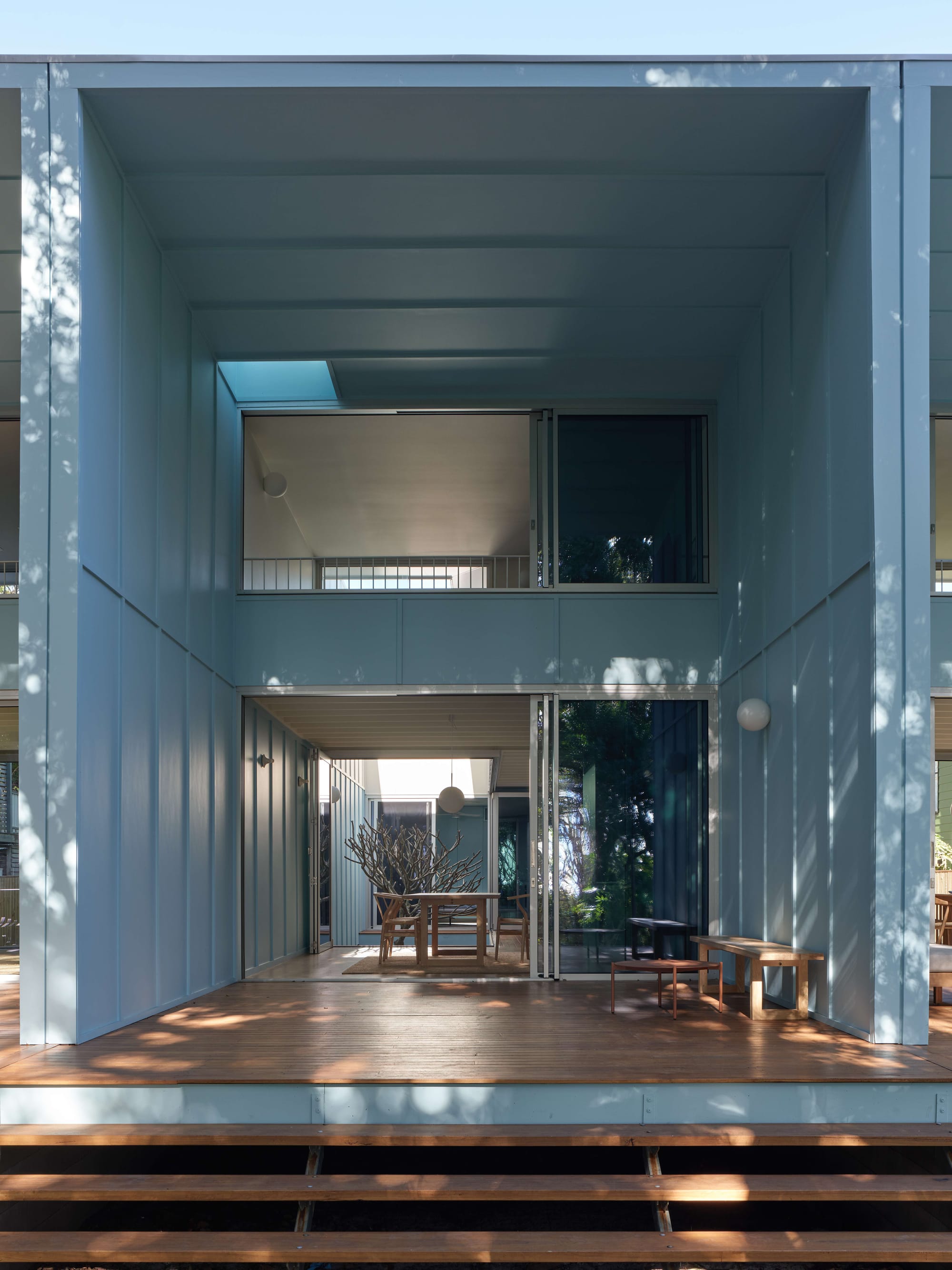 Blok Peregian by Blok Three Sisters. Photography by Christopher Frederick Jones. Rear facade of home with blue metal cladding, timber deck with stairs and full height glass sliding doors. 
