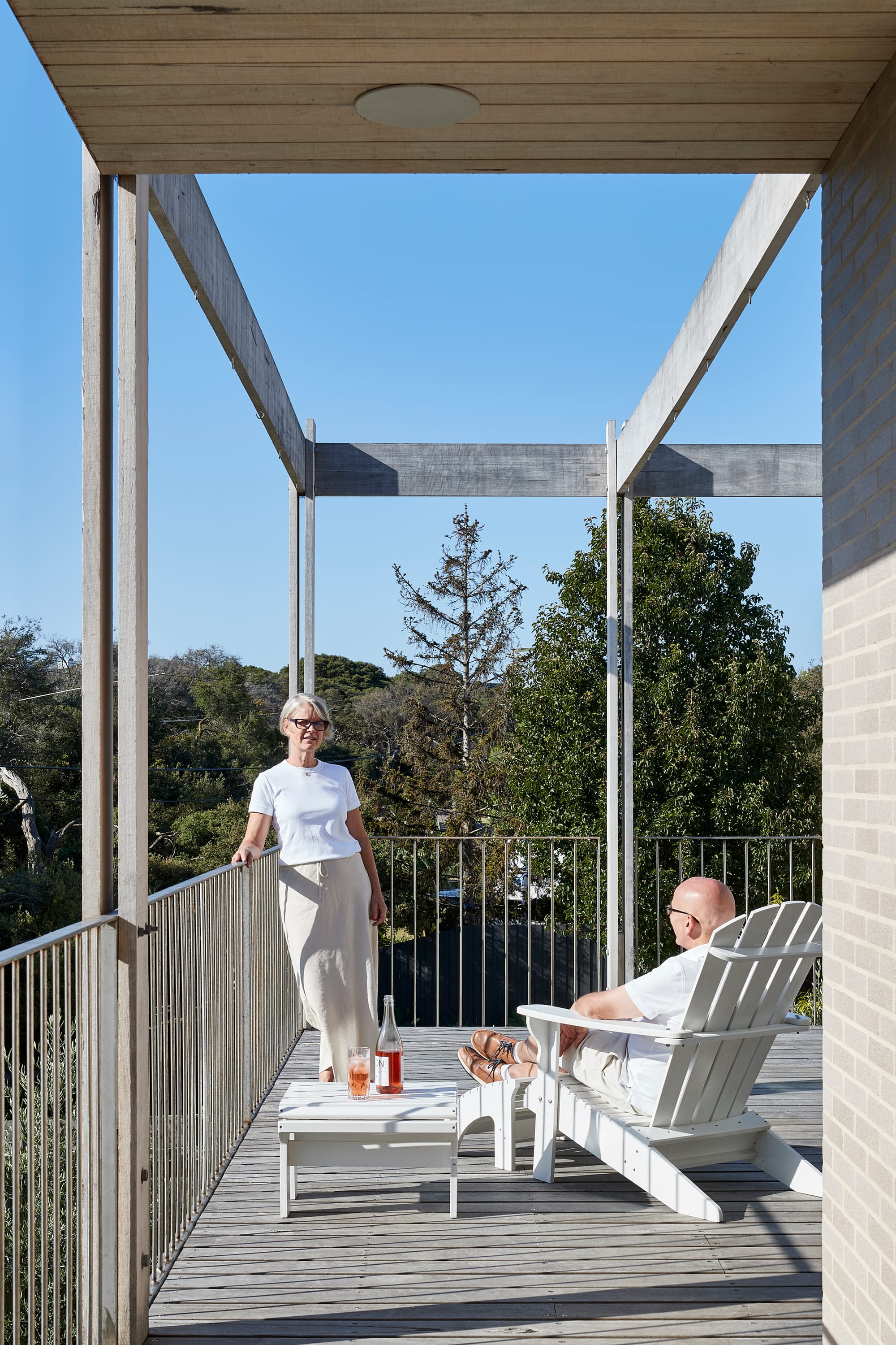The Blairgowrie Beach Club by Bower Architecture & Interiors. Photography by Shannon McGrath. Two people relaxing on a deck; one is seated and other on standing against metal railing wrapped around the deck.