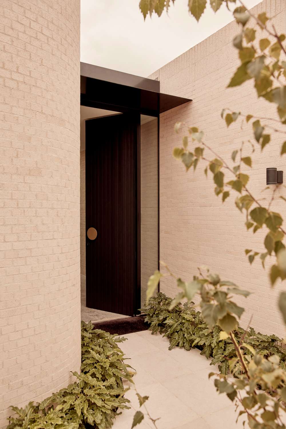 Brixton by Cera Stribley. Photography by Martina Gemmola. An entrance with curved pale brick walls, a dark vertical timber door with a round handle, black metal canopy, and surrounding greenery.