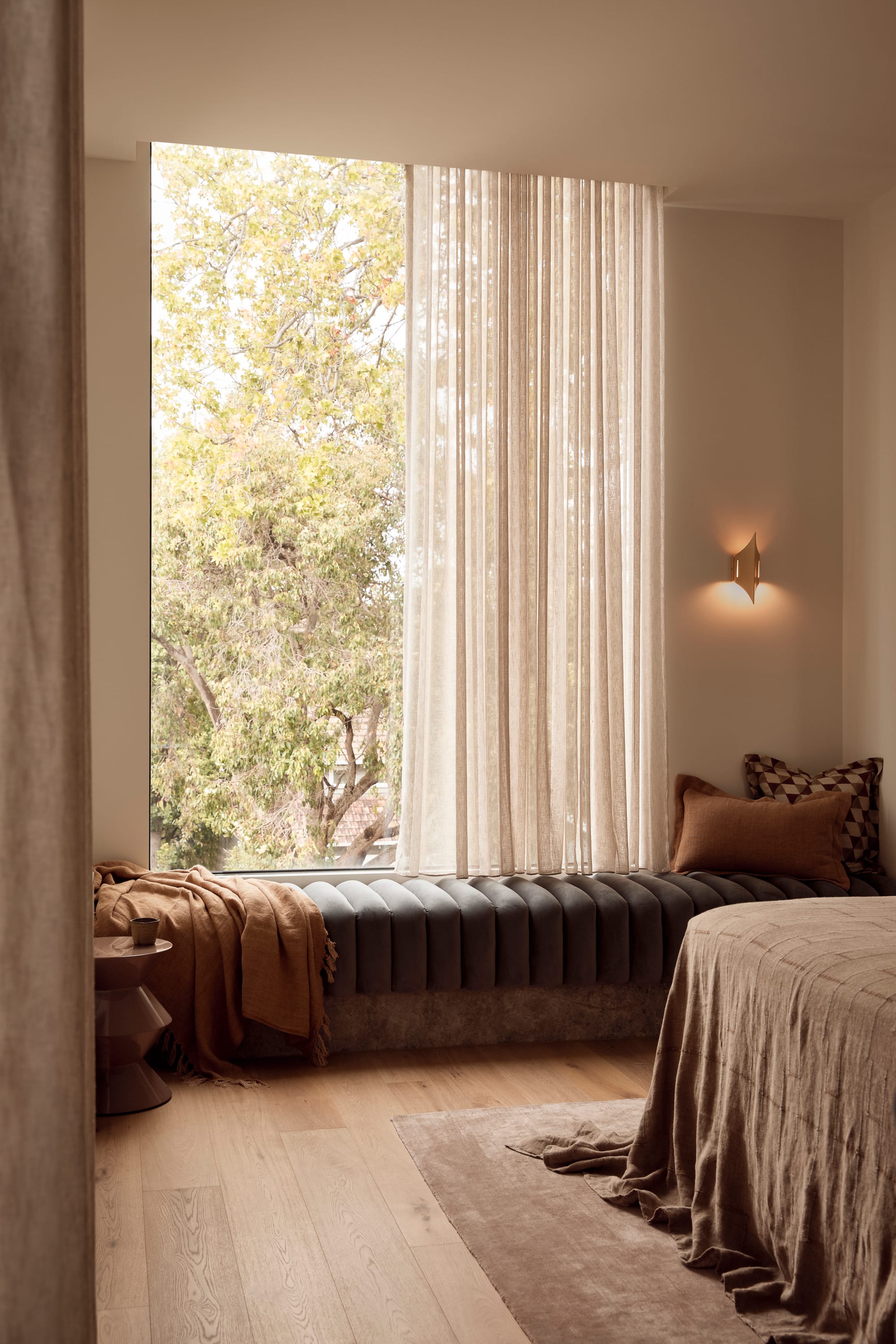 Brixton by Cera Stribley. Photography by Martina Gemmola. Warm, softly lit bedroom with a tall window seat, sheer curtains, earthy textiles, and a sculptural wall sconce.