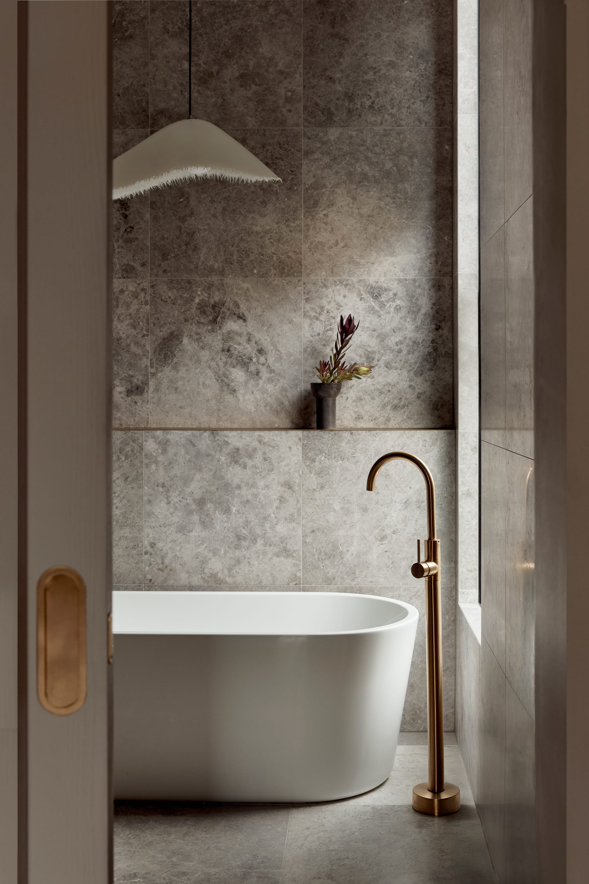 Brixton by Cera Stribley. Photography by Martina Gemmola. White bathtub with brass colored shower tap standing on side with grey tiles on the wall.