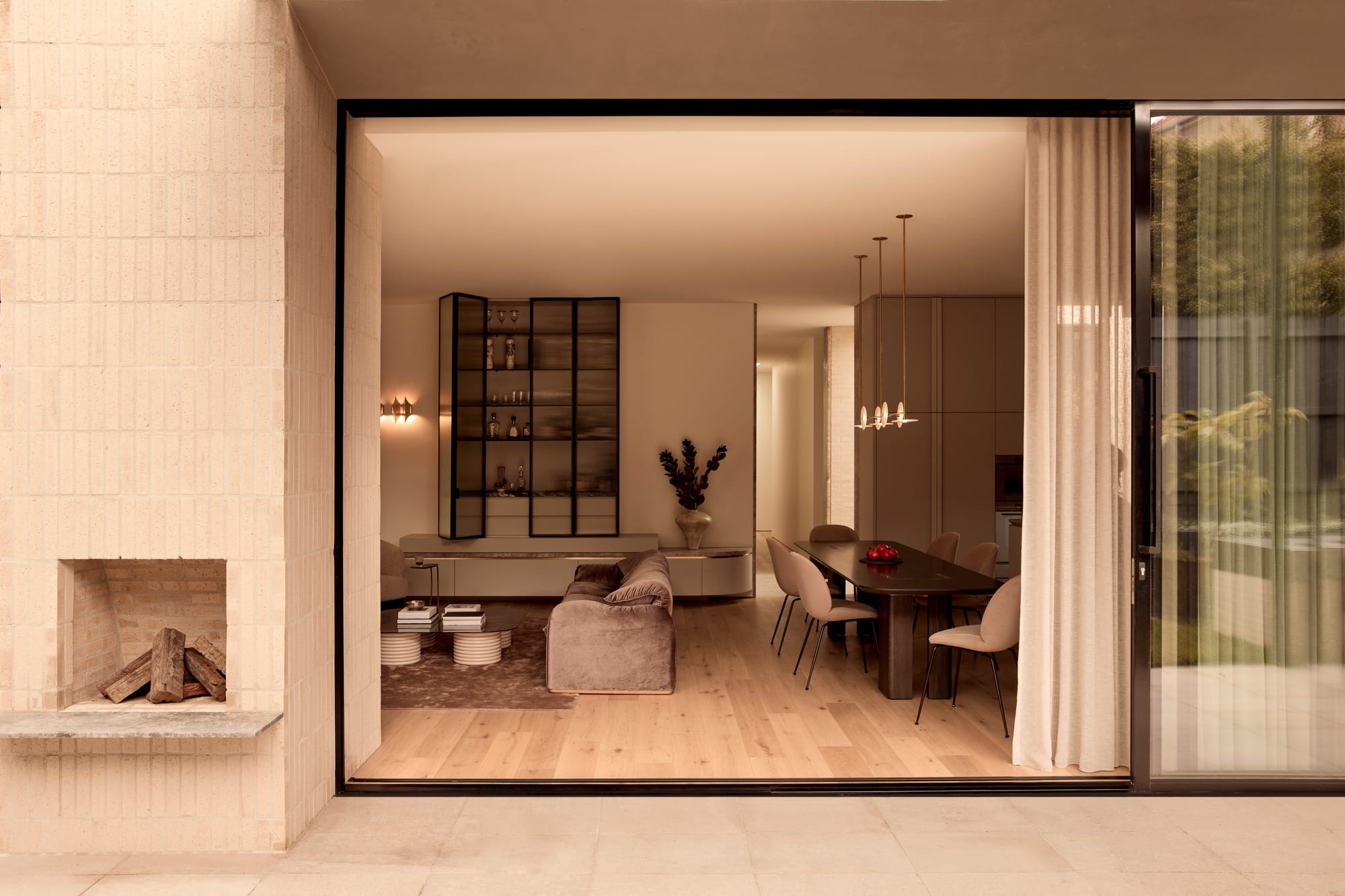 Brixton by Cera Stribley. Photography by Martina Gemmola. View into a refined open-plan living and dining space with soft neutral tones, a plush sofa, sleek black dining table, and glass cabinetry, framed by sliding doors and outdoor fireplace.