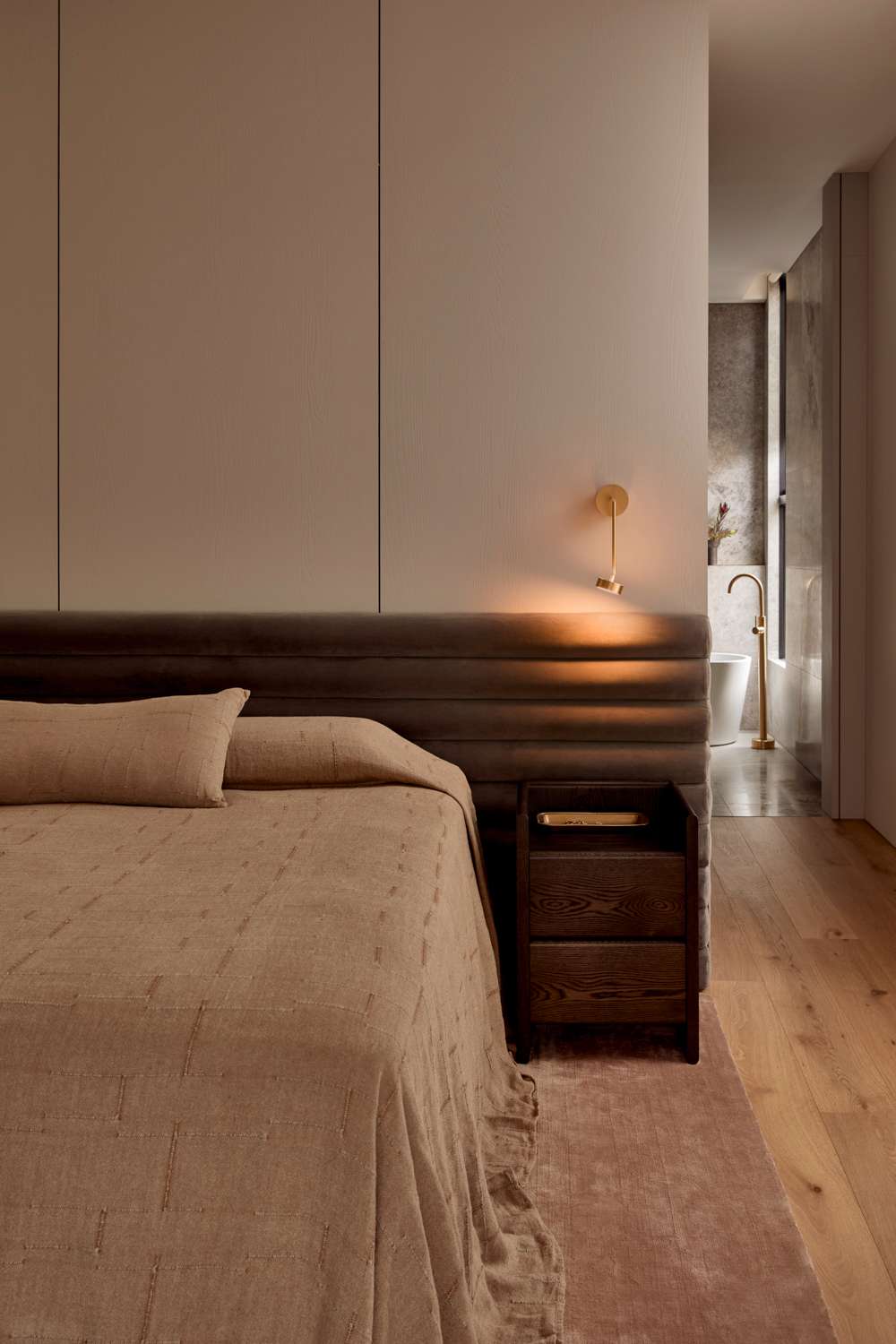 Brixton by Cera Stribley. Photography by Martina Gemmola. Bedroom with brown colored bedding with wooden side table and small bedside lamp hanging from the wall.