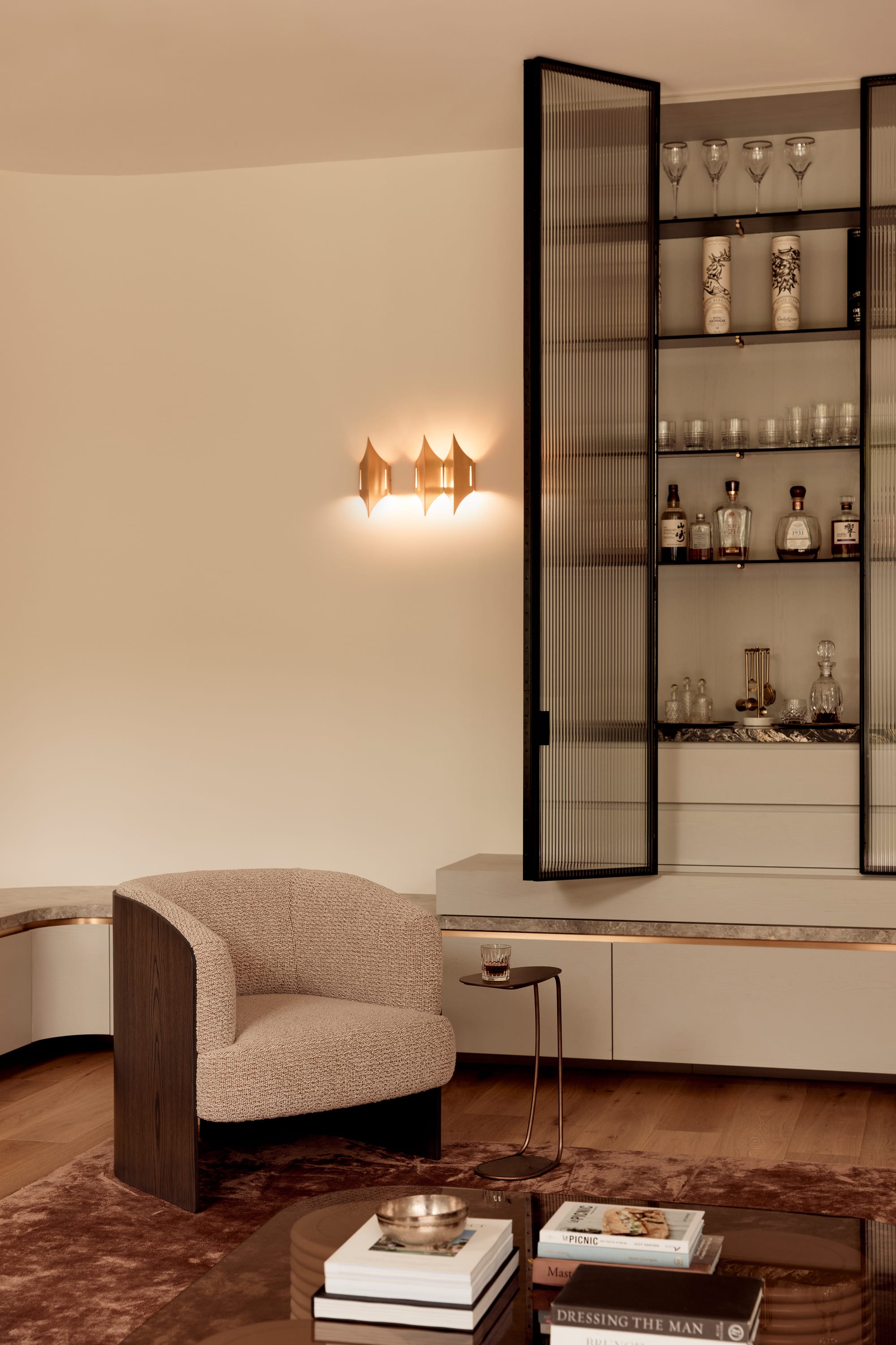 Brixton by Cera Stribley. Photography by Martina Gemmola. Elegant living space with a boucle armchair, sculptural wall sconce, glass-fronted bar cabinet, and a coffee table styled with books and brass decor.