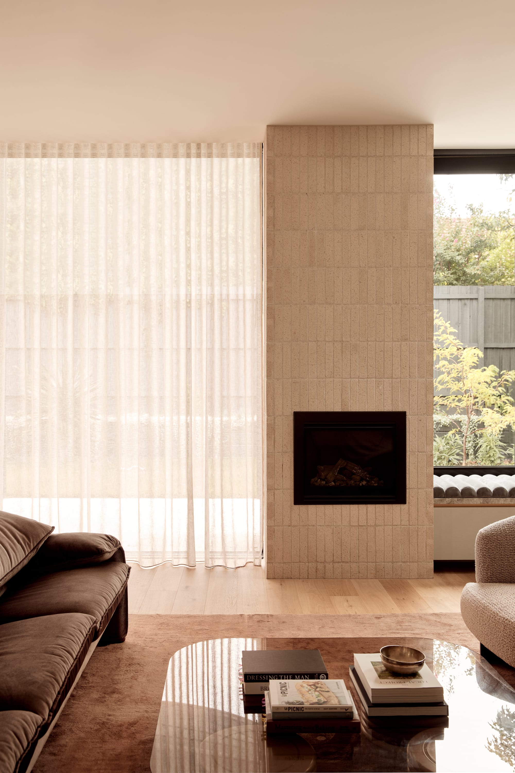 Brixton by Cera Stribley. Photography by Martina Gemmola. Tranquil living room with sheer curtains, light brick fireplace, velvet sofa, and coffee table stacked with books in a soft, neutral palette.