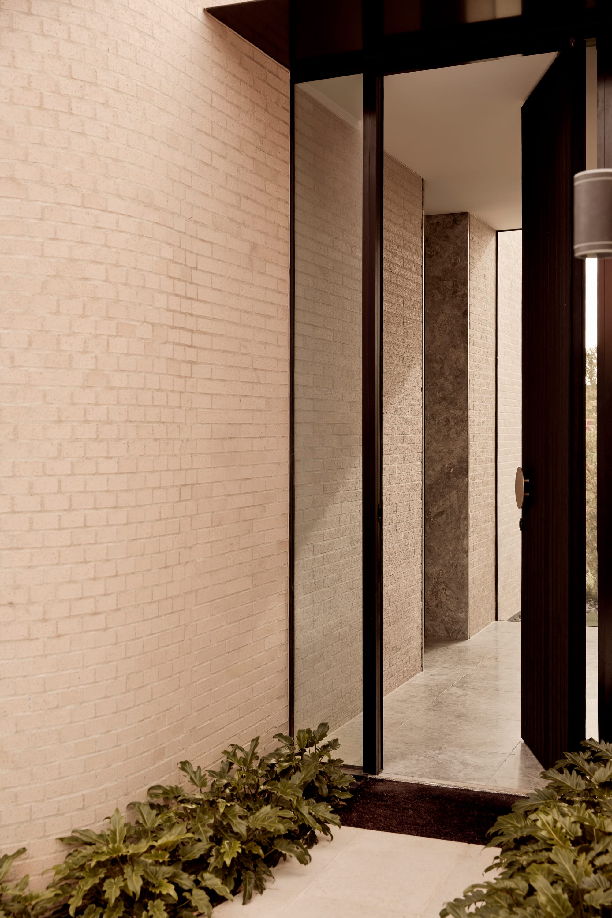 Brixton by Cera Stribley. Photography by Martina Gemmola. Refined entryway with tall pivot door, pale brick walls, polished stone flooring, and soft landscaping flanking the threshold.