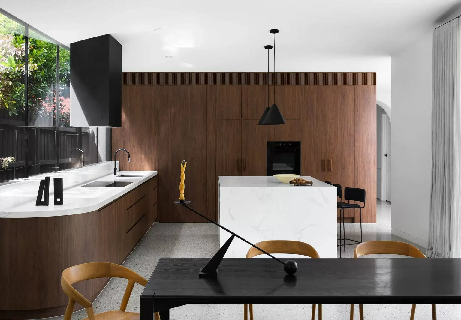 Maple House by Cera Stribley. Photography by Emily Bartlett. Kitchen with curved timber bench, white stone island bench and countertops, concrete floors and black details.  