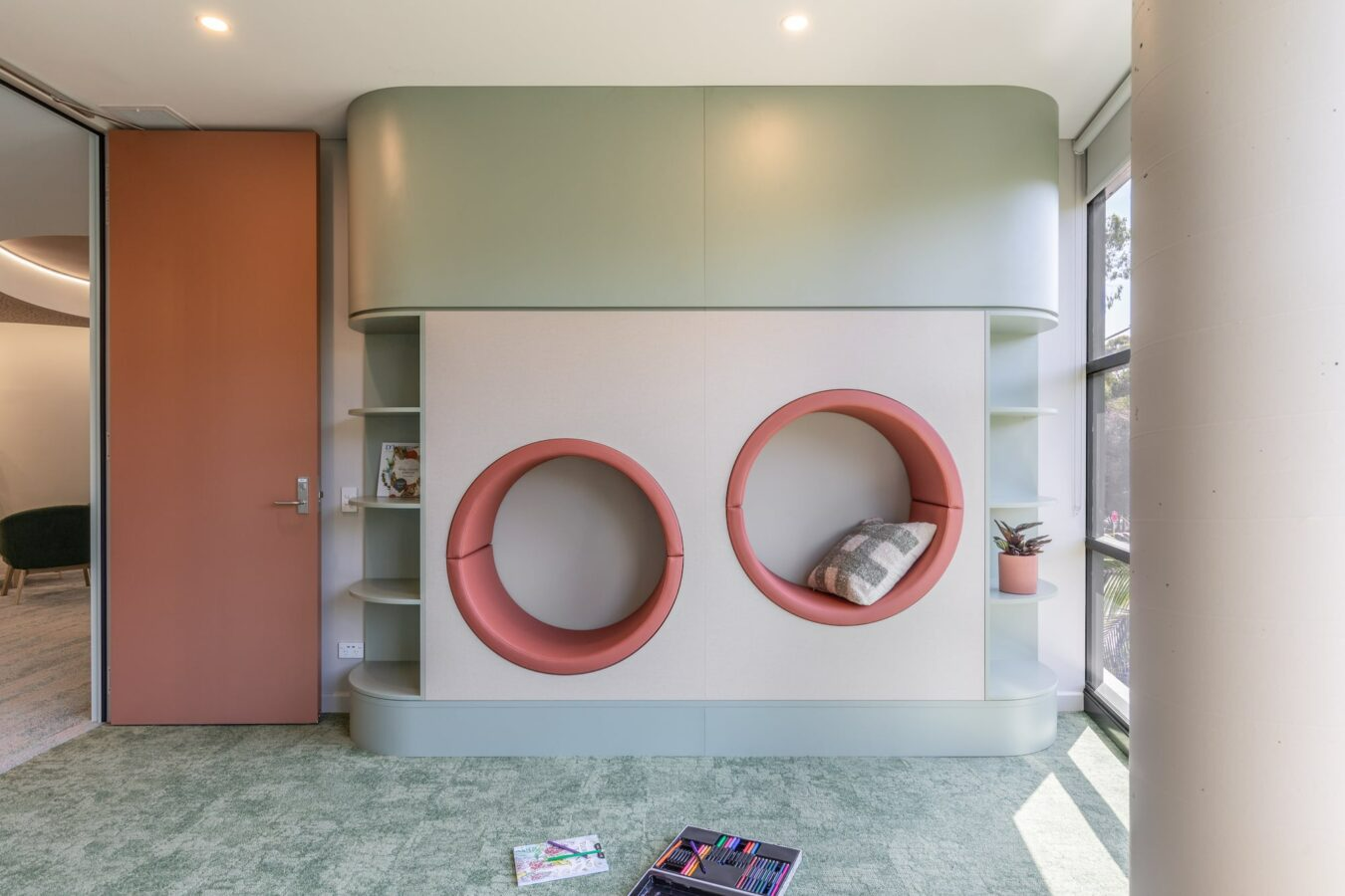 Women’s Trauma Recovery Centre & Health Consulting Rooms by Edmiston Jones. Photographer undisclosed. Integrated wall seating and storage with mint panelling and coral curved seat cushions. 