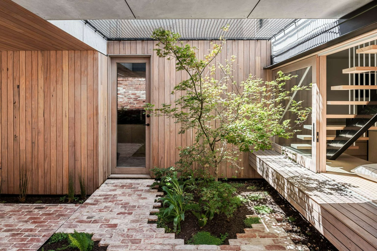 Pop-Up House by FIGR Architecture Studio. Photography by Tom Blachford. Interior courtyard space wwith timber clad walls and decking, reclaimed brick flooring, a tree in a garden bed and a net ceiling.