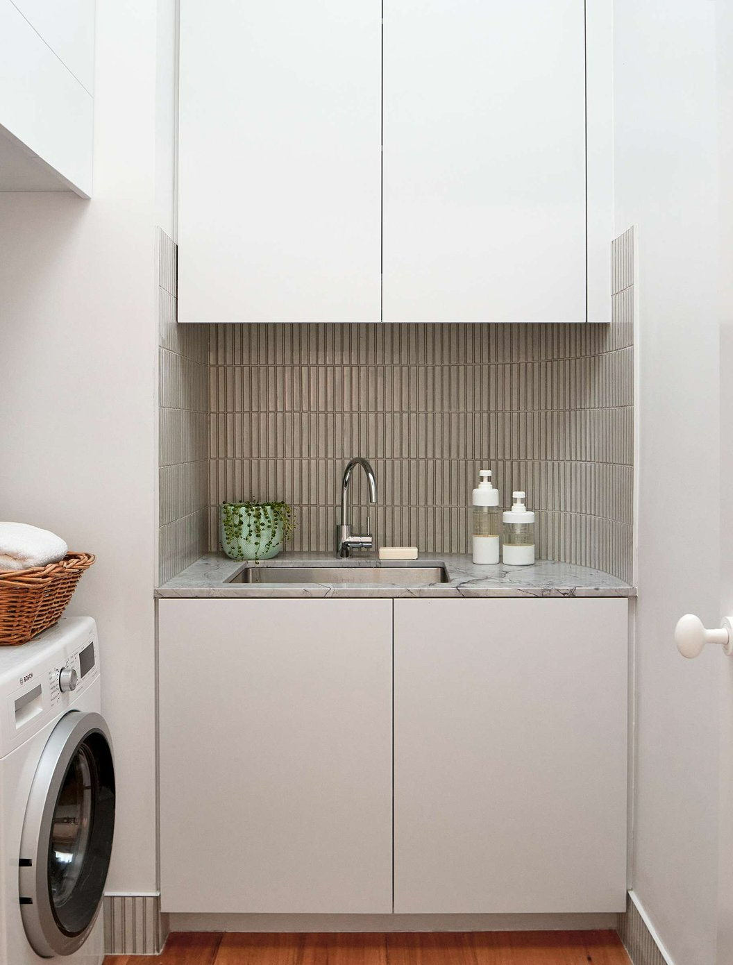 Envelop House by mcmahon and nerlich. Photography by Shannon McGrath. Small laundry space with hardwood timber floors, white cabinets, marble benchtops beige finger tile splashbacks and overhead white cupboards. 