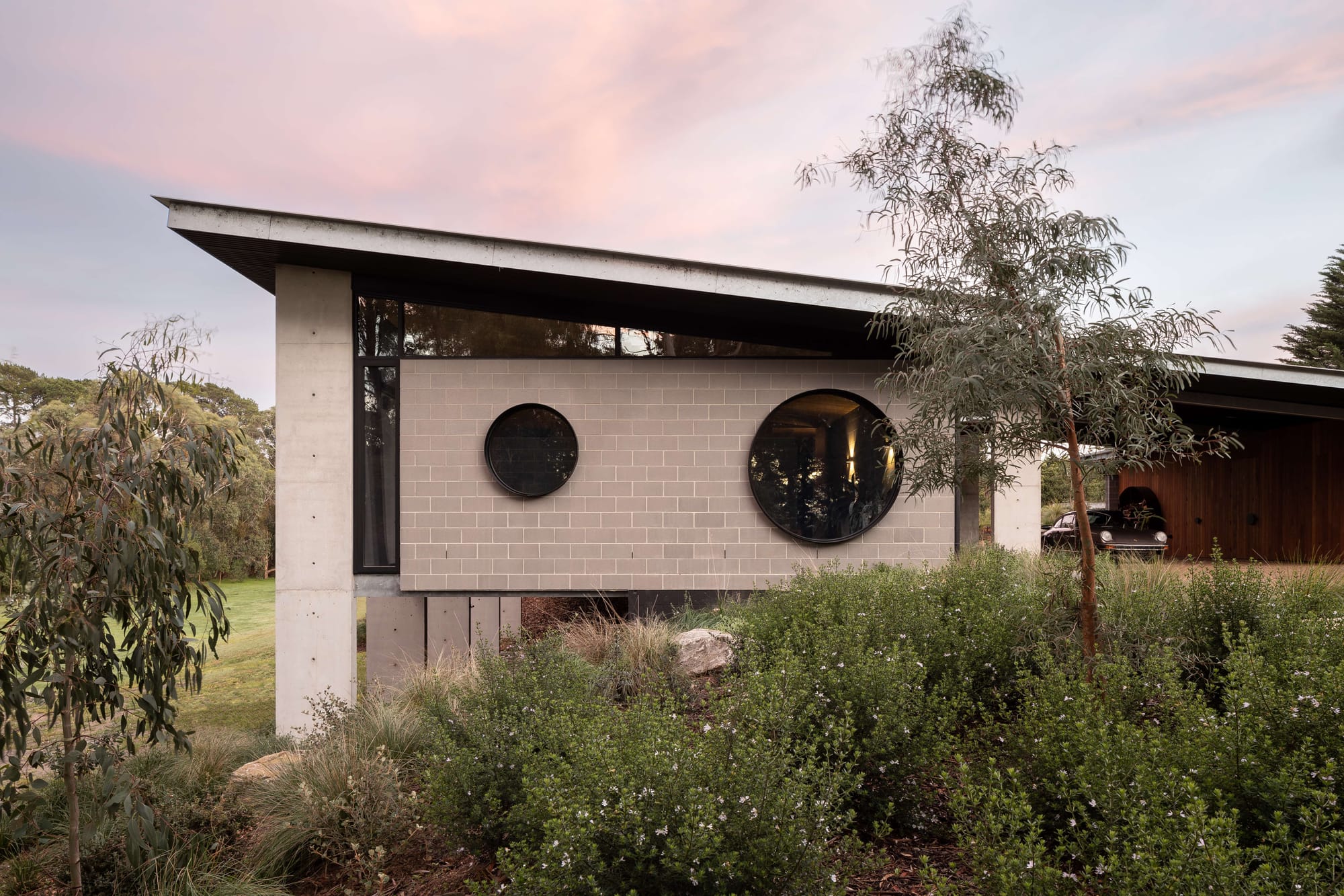 Merricks House by Aktis Architects. Photography by&nbsp;Timothy Kaye. Modern house with angled roof, concrete and blockwork walls, and two circular windows set against a lush native garden at dusk.