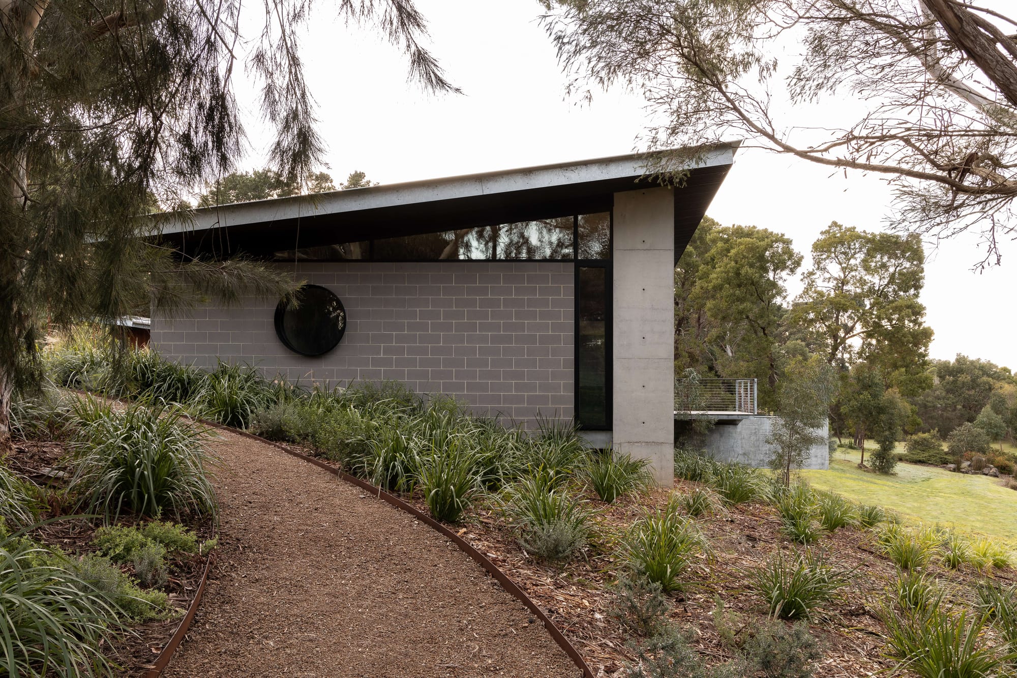 Merricks House by Aktis Architects. Photography by&nbsp;Timothy Kaye. Side view of a modern home with circular window, grey blockwork, and angled roof, bordered by a winding gravel path and native landscaping.