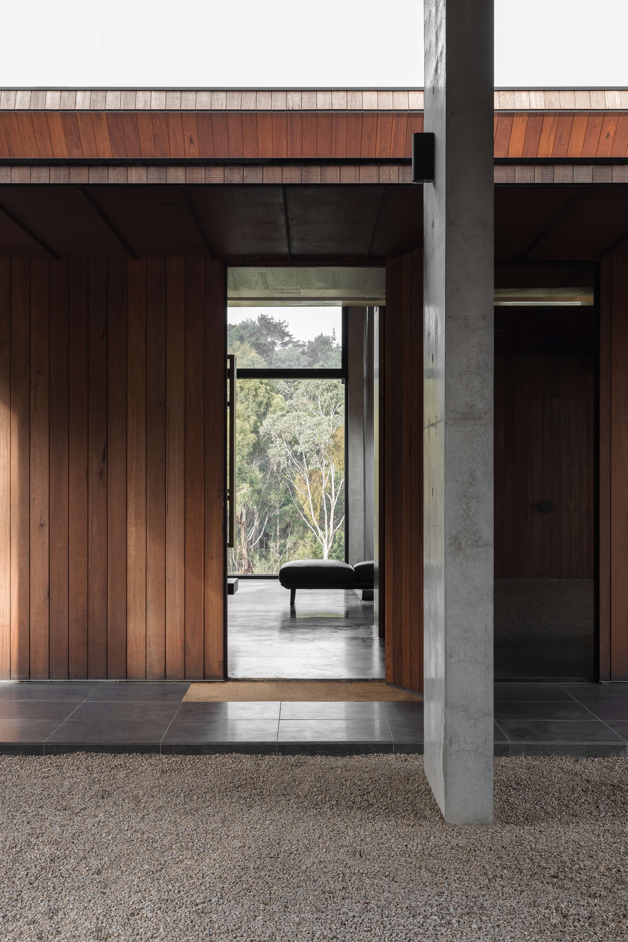 Merricks House by Aktis Architects. Photography by&nbsp;Timothy Kaye.Framed view through timber-clad entry and concrete column to a bench seat and forest beyond, with gravel and tiled flooring.