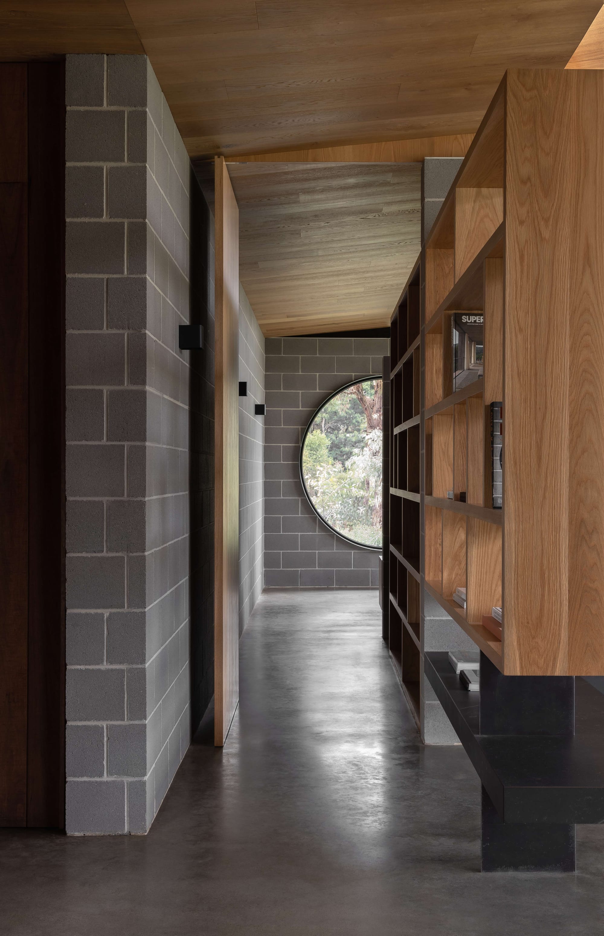 Merricks House by Aktis Architects. Photography by&nbsp;Timothy Kaye. Interior hallway with concrete block walls, timber ceiling, floating bookshelves, and a large circular window framing a garden view.