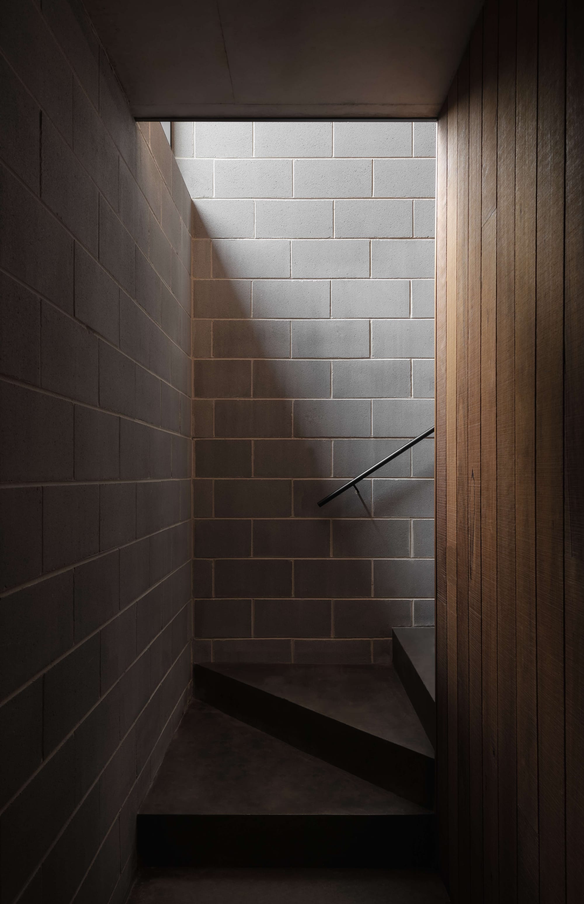 Merricks House by Aktis Architects. Photography by&nbsp;Timothy Kaye. Minimal stairwell with grey concrete block walls, timber cladding, and soft natural light casting shadows across the textured surfaces.