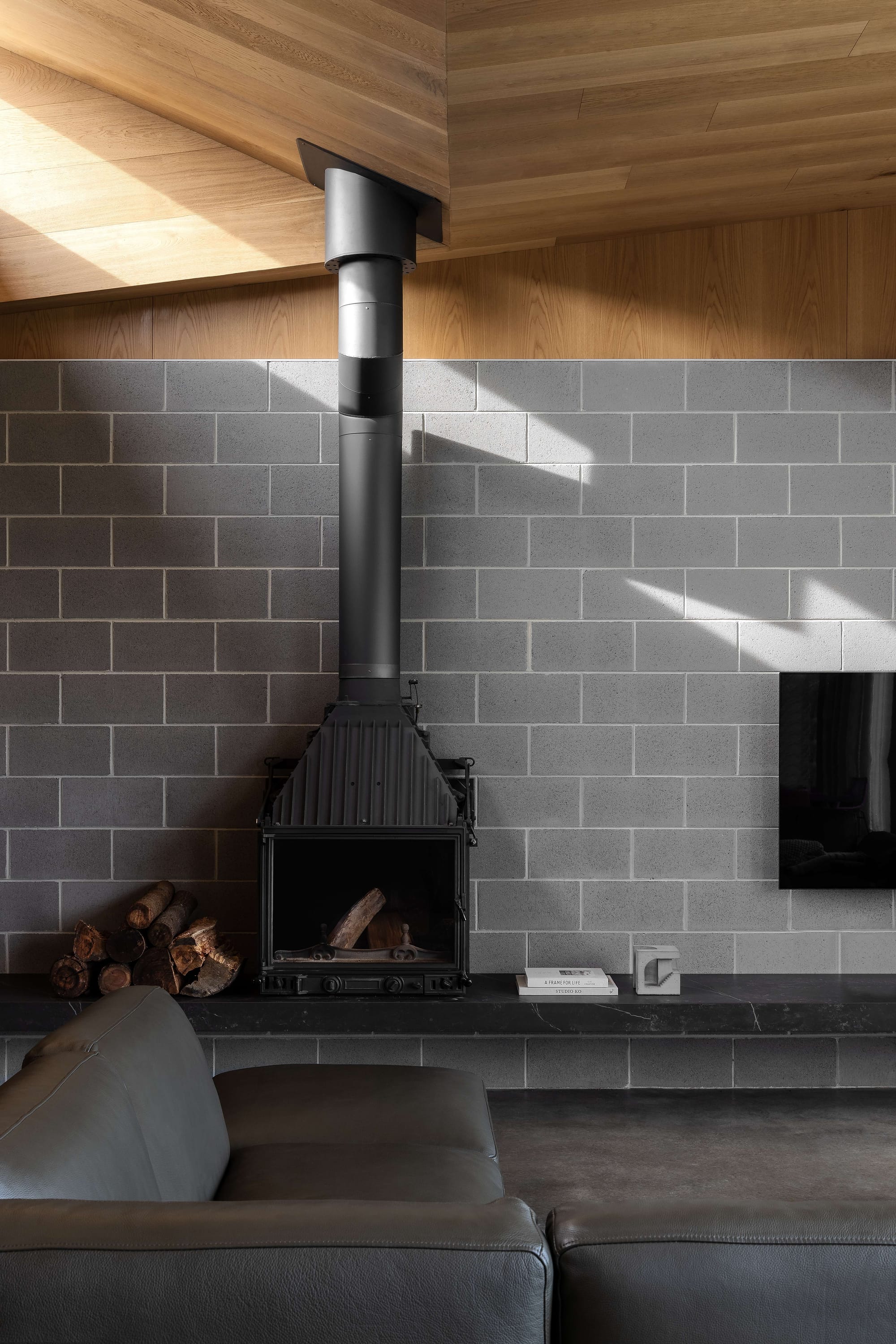 Merricks House by Aktis Architects. Photography by&nbsp;Timothy Kaye. Close-up of a modern wood-burning fireplace with a black flue, set against a grey blockwork wall and timber ceiling, with stacked firewood and minimalist decor.