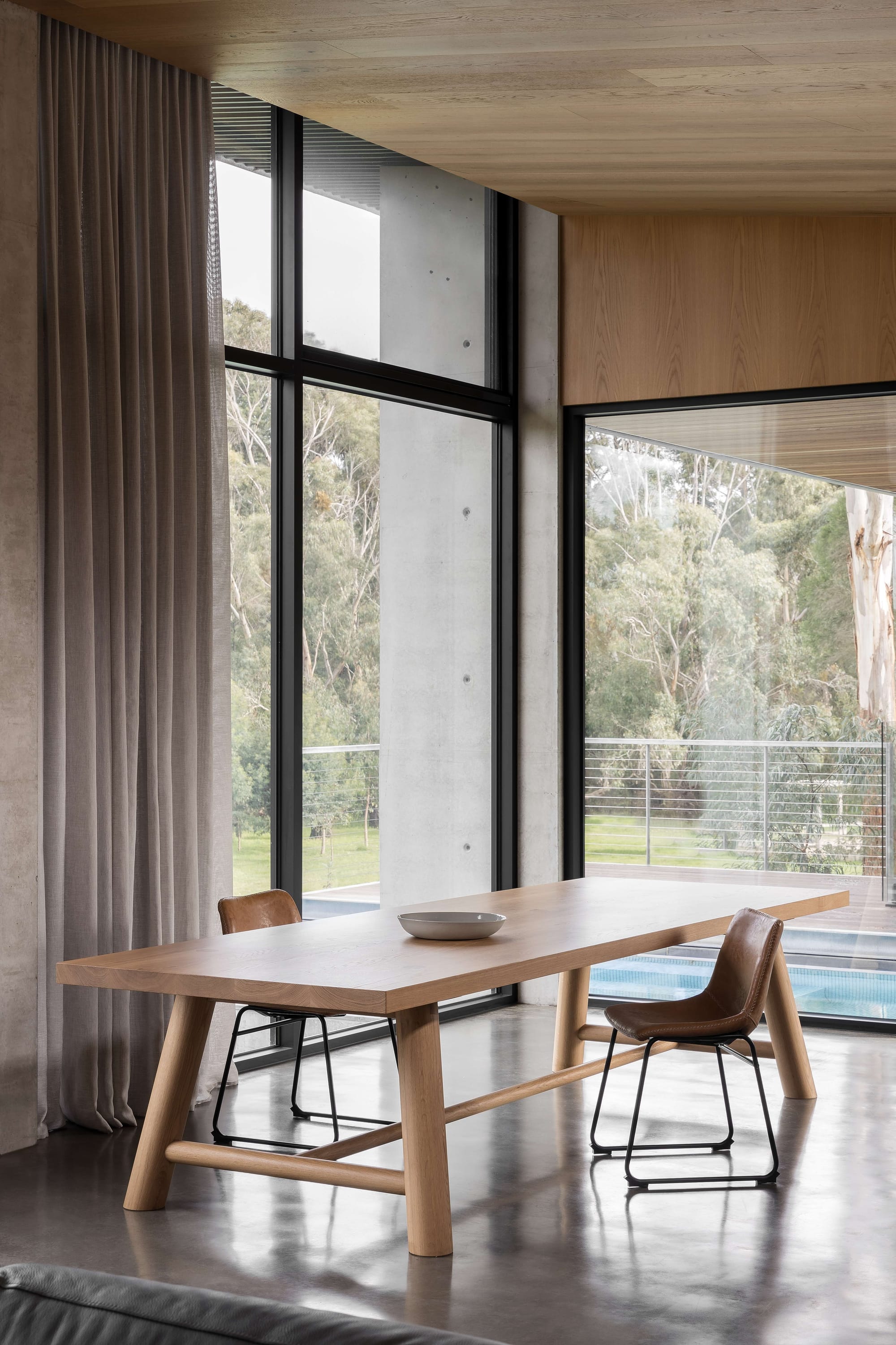 Merricks House by Aktis Architects. Photography by&nbsp;Timothy Kaye. Dining area with a solid timber table, leather chairs, full-height windows, and views to a landscaped garden and pool beyond.