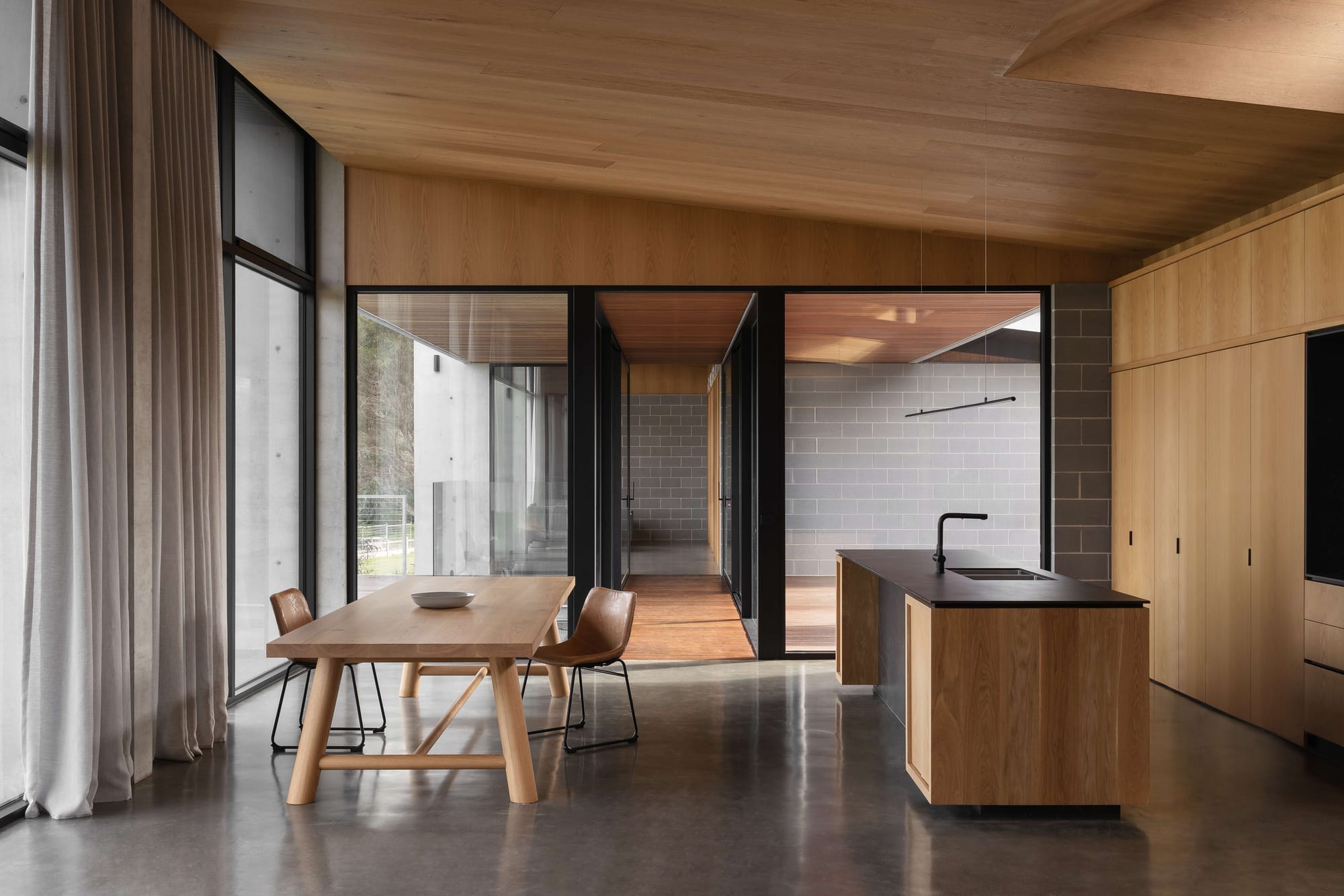 Merricks House by Aktis Architects. Photography by&nbsp;Timothy Kaye. Open-plan kitchen and dining area with timber cabinetry, black benchtop, large glass doors, and polished concrete flooring.