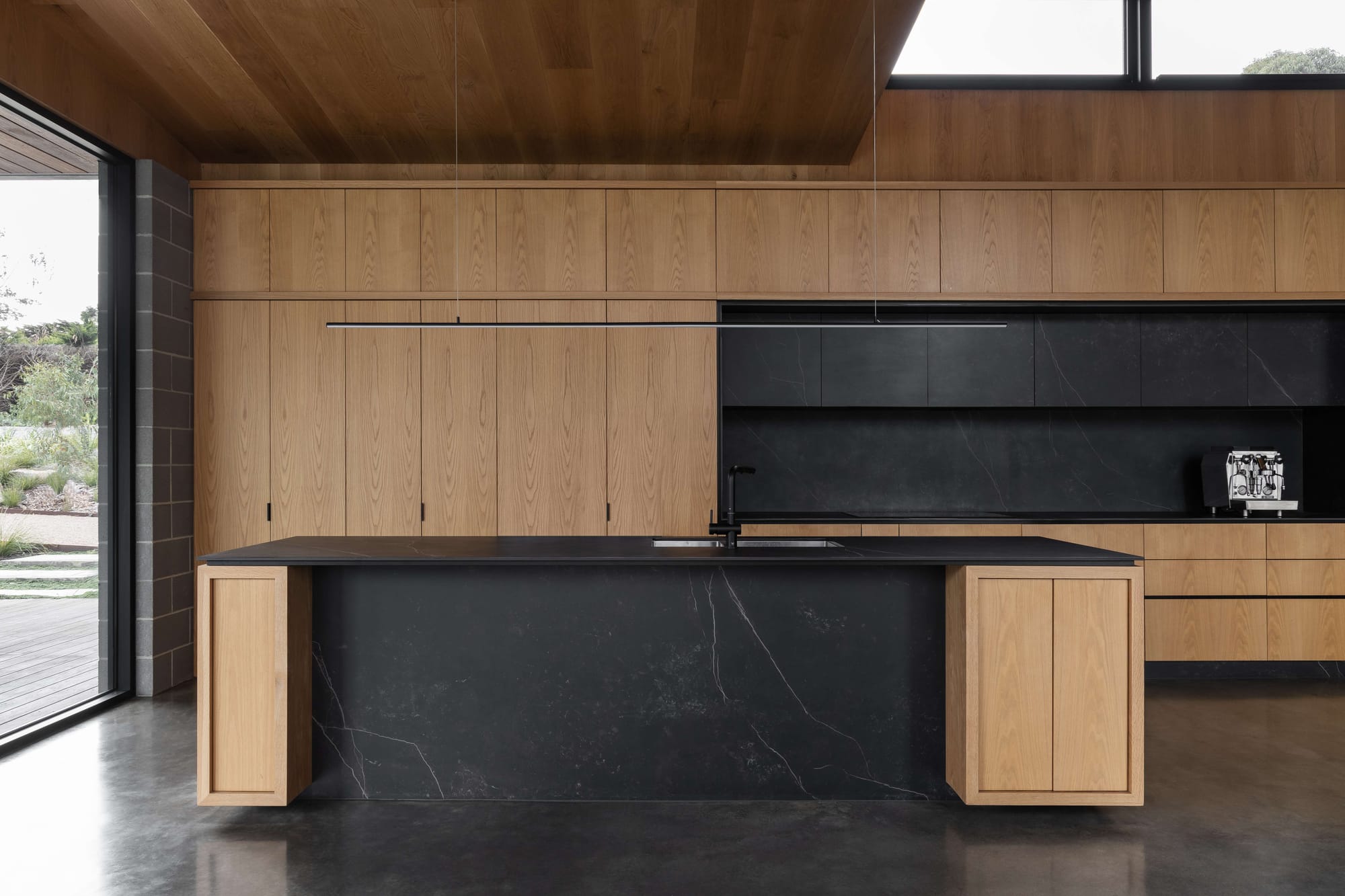Merricks House by Aktis Architects. Photography by&nbsp;Timothy Kaye. Contemporary kitchen with black marble island, timber cabinetry, minimalist pendant lighting, and polished concrete floors.