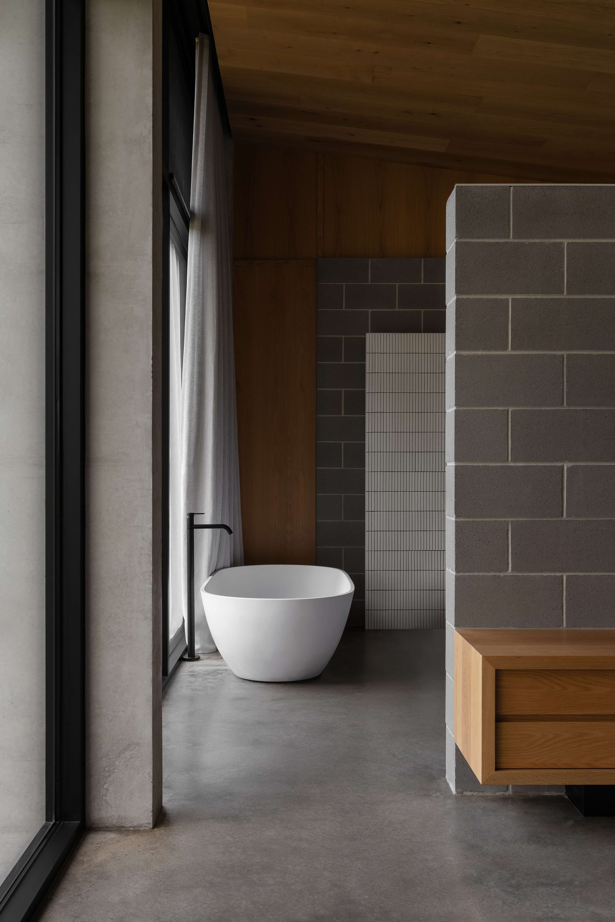 Merricks House by Aktis Architects. Photography by&nbsp;Timothy Kaye.  Serene bathroom with freestanding white tub, grey concrete block walls, minimalist black tapware, and full-height windows with sheer curtains.