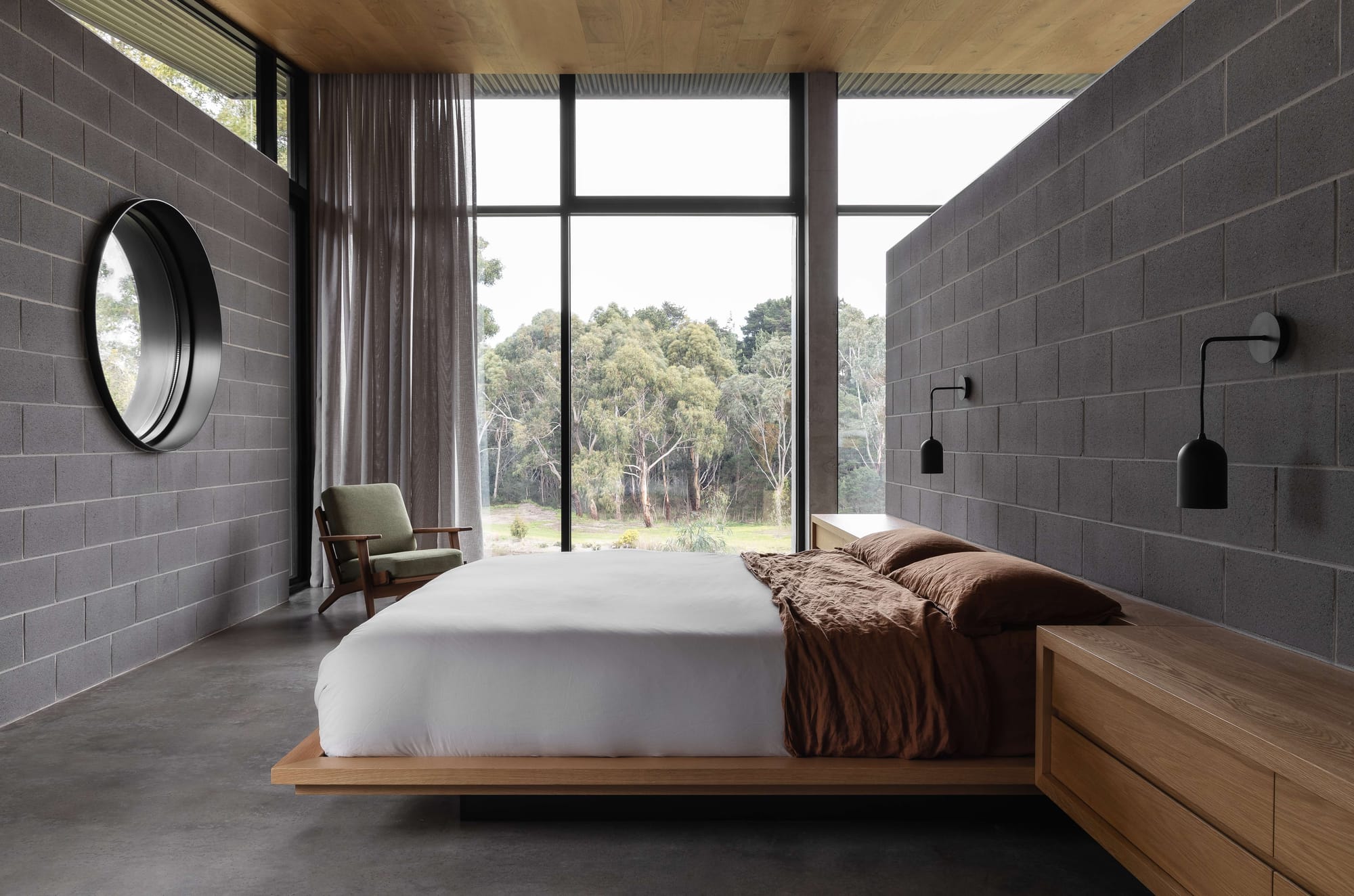 Merricks House by Aktis Architects. Photography by&nbsp;Timothy Kaye. Minimalist bedroom with concrete block walls, timber floating platform bed, large picture window framing forest views, and a circular wall mirror.