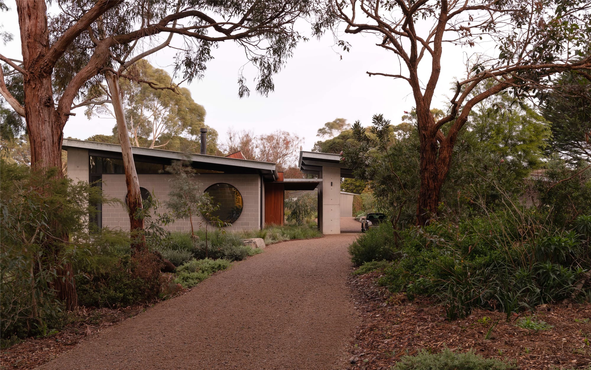 Merricks House by Aktis Architects. Photography by&nbsp;Timothy Kaye. Exterior view of a modern house nestled in native Australian bushland, featuring grey blockwork walls, a distinctive round window, and a low-sloped metal roof surrounded by tall eucalyptus trees.