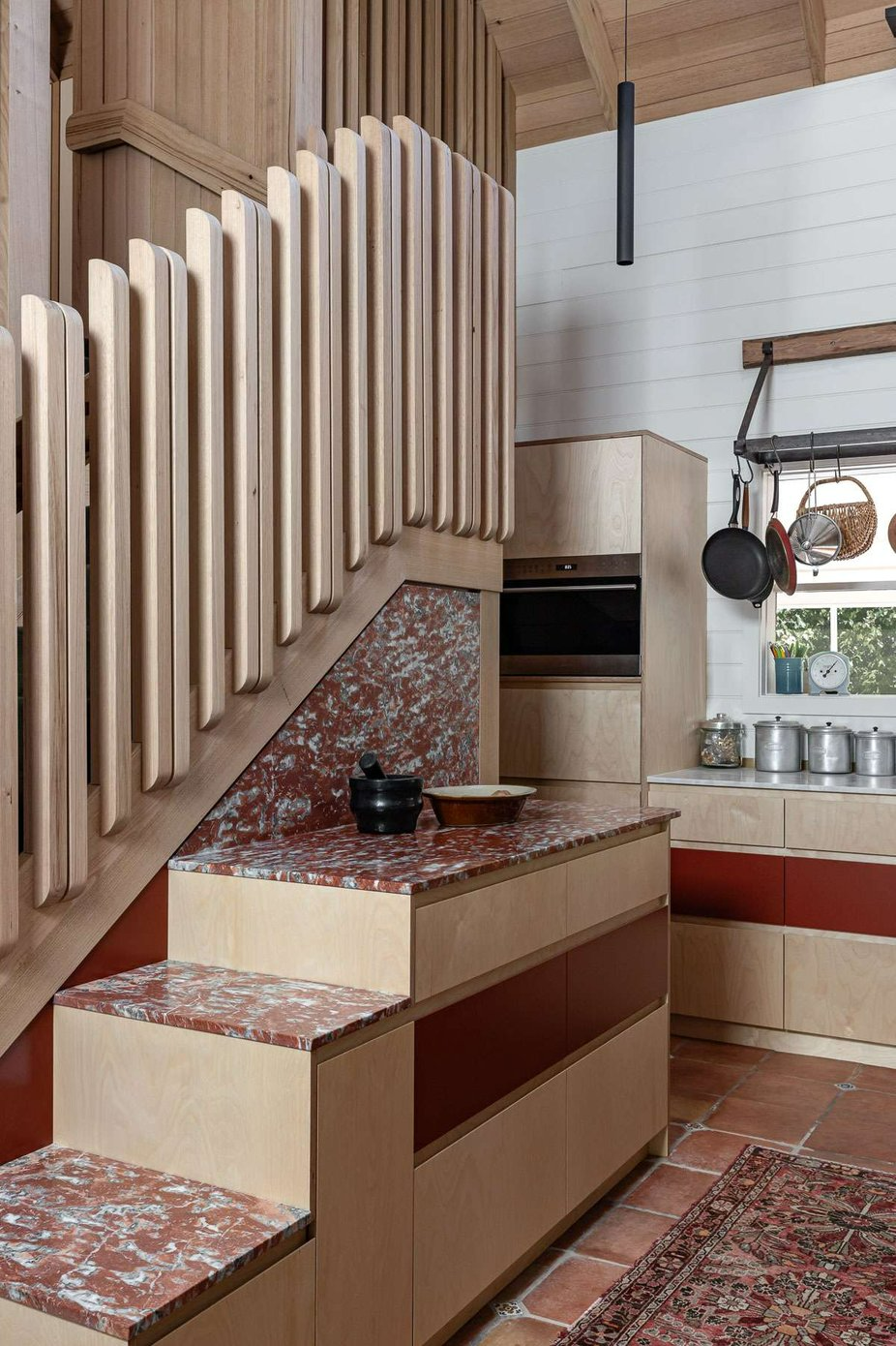 Redbarn by Roberston Collectif. Photography by Life In Still. Staggered bench and storage with red marble benchtop, terracotta floor tiles and maroon and plywood cabinet faces. 