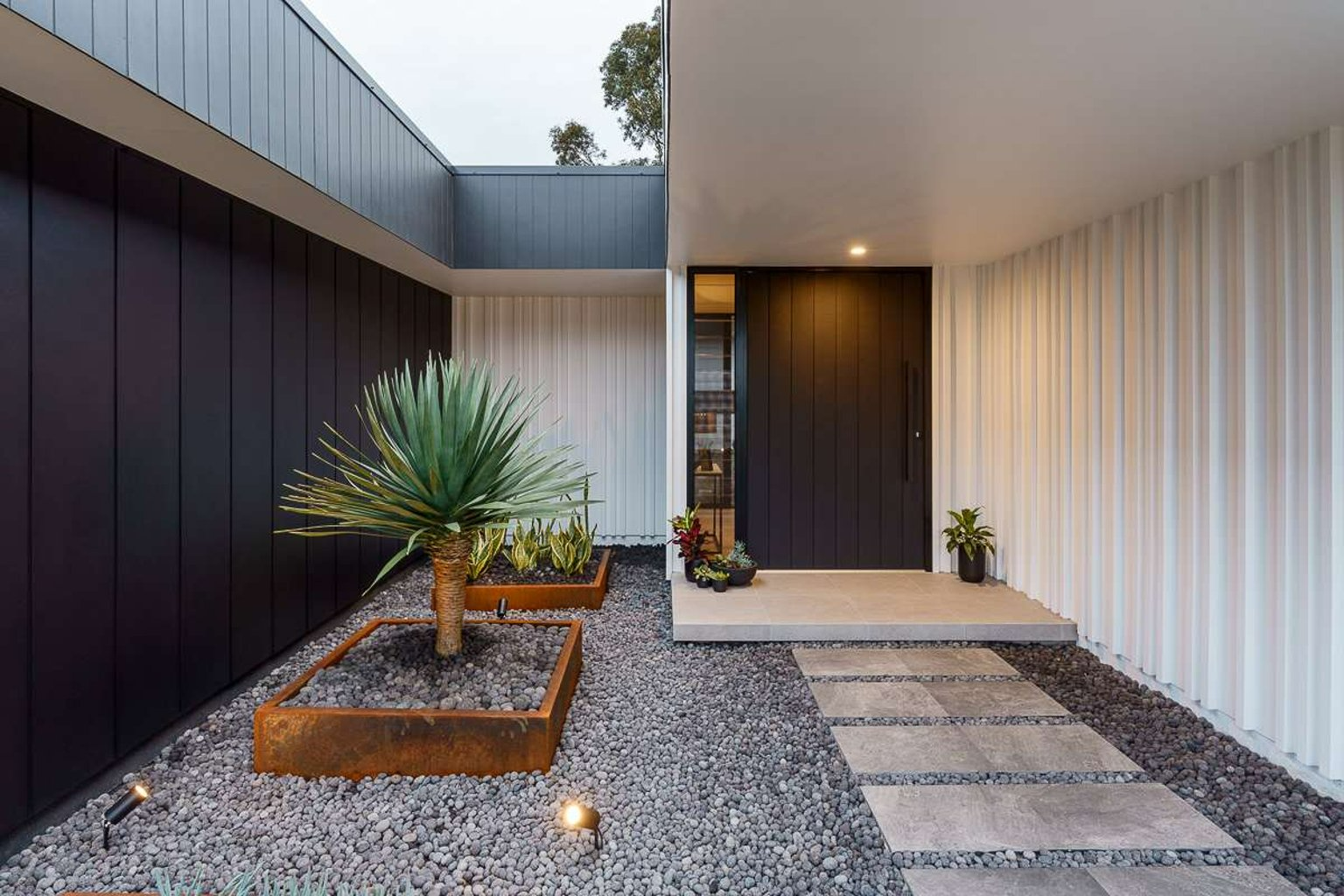 Hunter Valley House by Sarah Waller Architecture. Photography by Joshua Hogan. Front entrance with pavers leading to door and weathered metal garden beds with agave plant. 