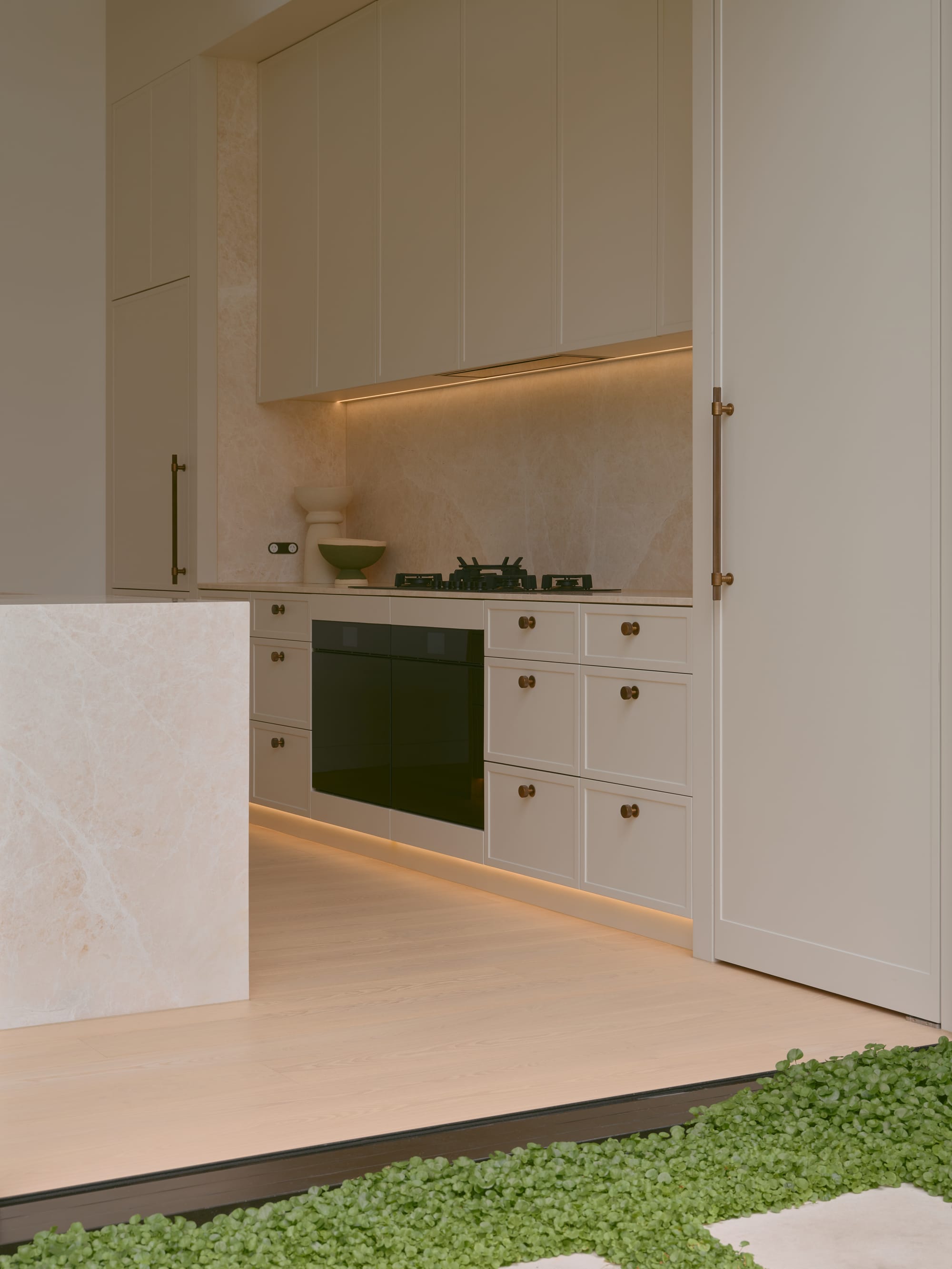 Sole House by 868 Architects. Photography by Martin Siegner. Detail view of a modern kitchen with matte cabinetry, bronze hardware, marble finishes, under-cabinet lighting, and seamless transition to an outdoor green space.