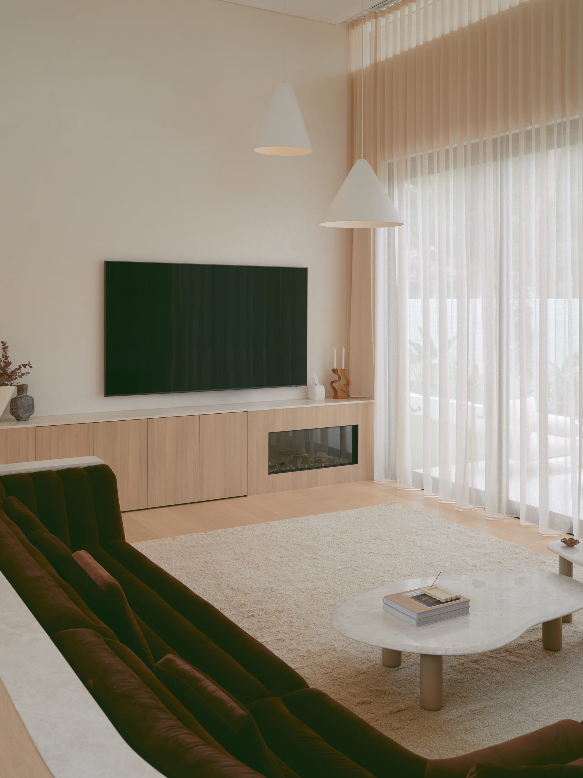 Sole House by 868 Architects. Photography by Martin Siegner. Minimalist living room with a velvet sofa, wall-mounted TV, light wood cabinetry, cone pendant lights, and sheer floor-to-ceiling curtains.