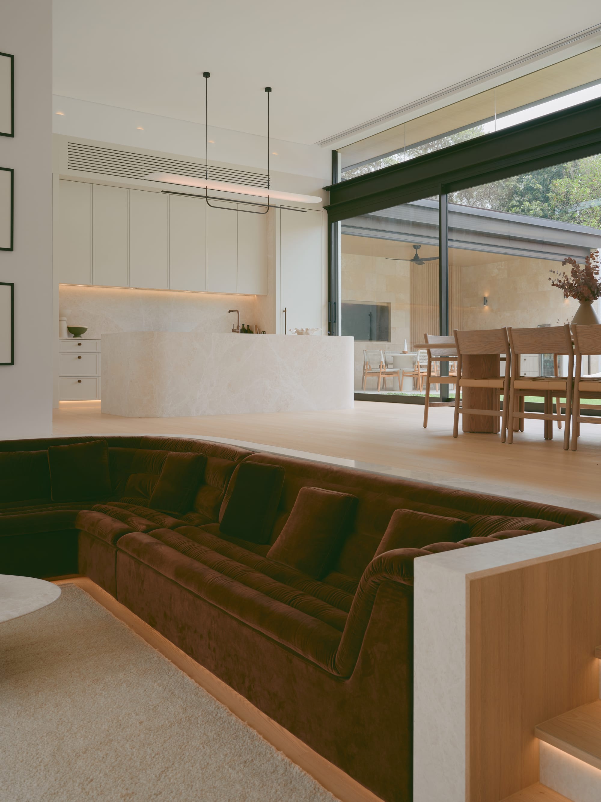 Sole House by 868 Architects. Photography by Martin Siegner. Open-plan living space featuring a sunken velvet lounge, minimalist marble kitchen, timber dining set, and floor-to-ceiling glass doors that connect to an outdoor area.