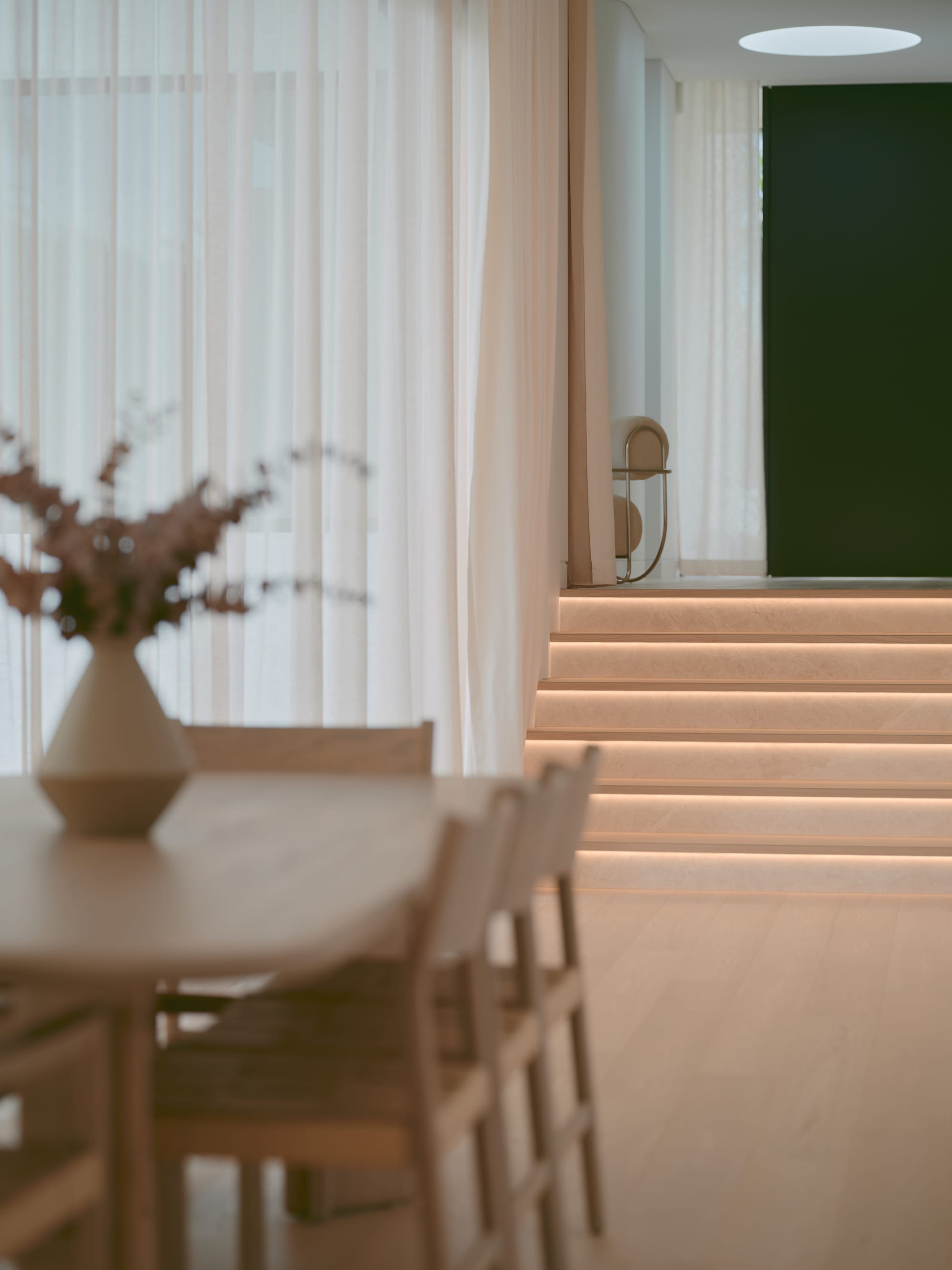 Sole House by 868 Architects. Photography by Martin Siegner. Close-up of a minimalist kitchen and dining area featuring light-toned timber furniture, a sculpted marble island, soft under-cabinet lighting, and sheer curtains that filter natural light.