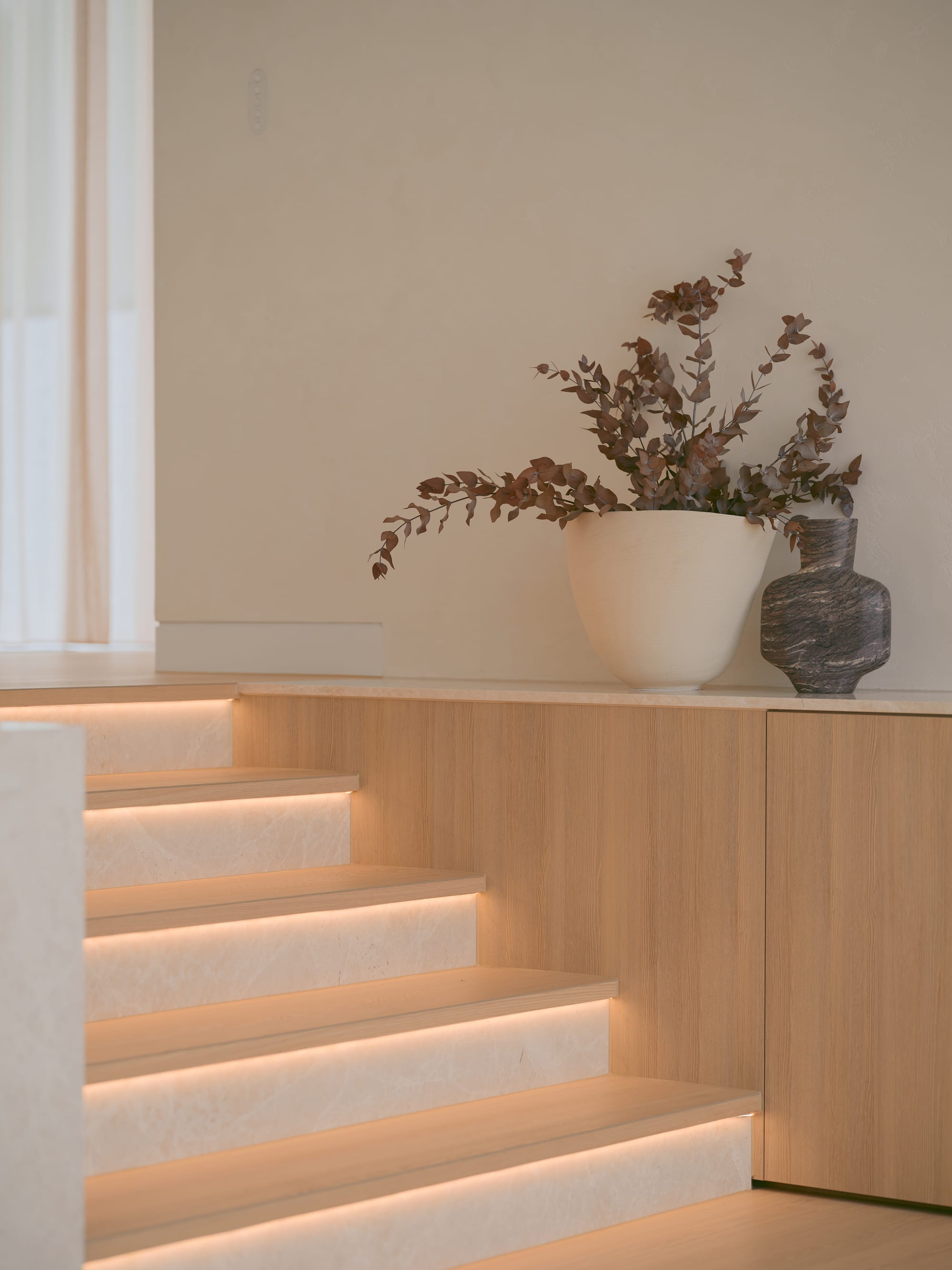 Sole House by 868 Architects. Photography by Martin Siegner. Softly illuminated timber and stone staircase with integrated lighting, flanked by minimalist cabinetry and styled with sculptural vases and dried foliage.