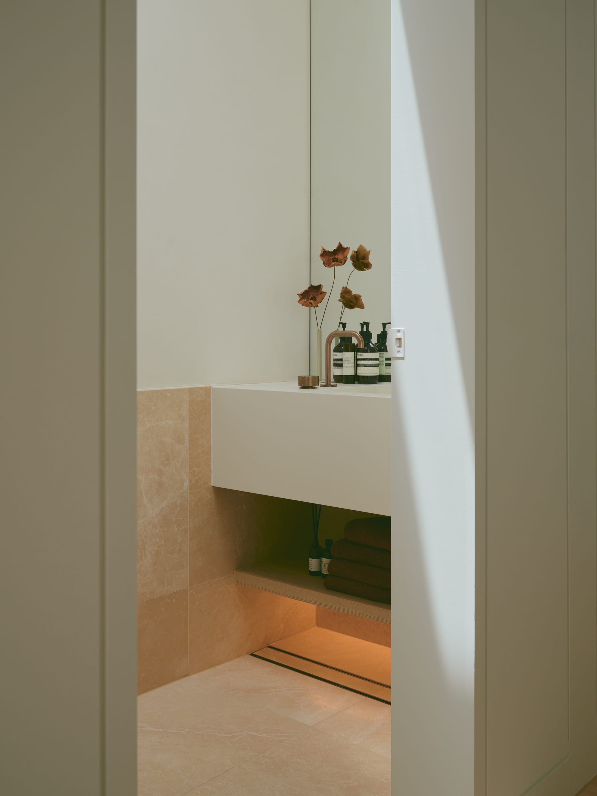 Sole House by 868 Architects. Photography by Martin Siegner. Minimal powder room with warm stone tiles, a floating vanity, integrated mirror, bronze tapware, and soft under-shelf lighting creating a tranquil ambiance.