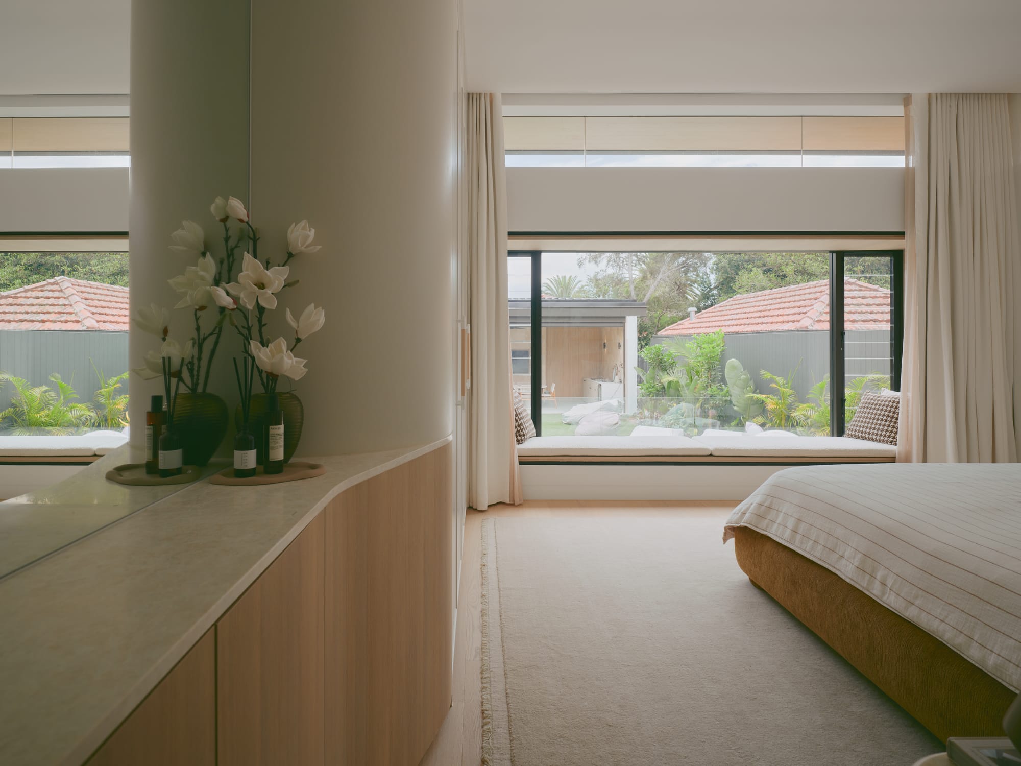 Sole House by 868 Architects. Photography by Martin Siegner. Serene bedroom with a curved feature wall, built-in cabinetry, window bench seating, and expansive glazing that opens to a lush garden view.