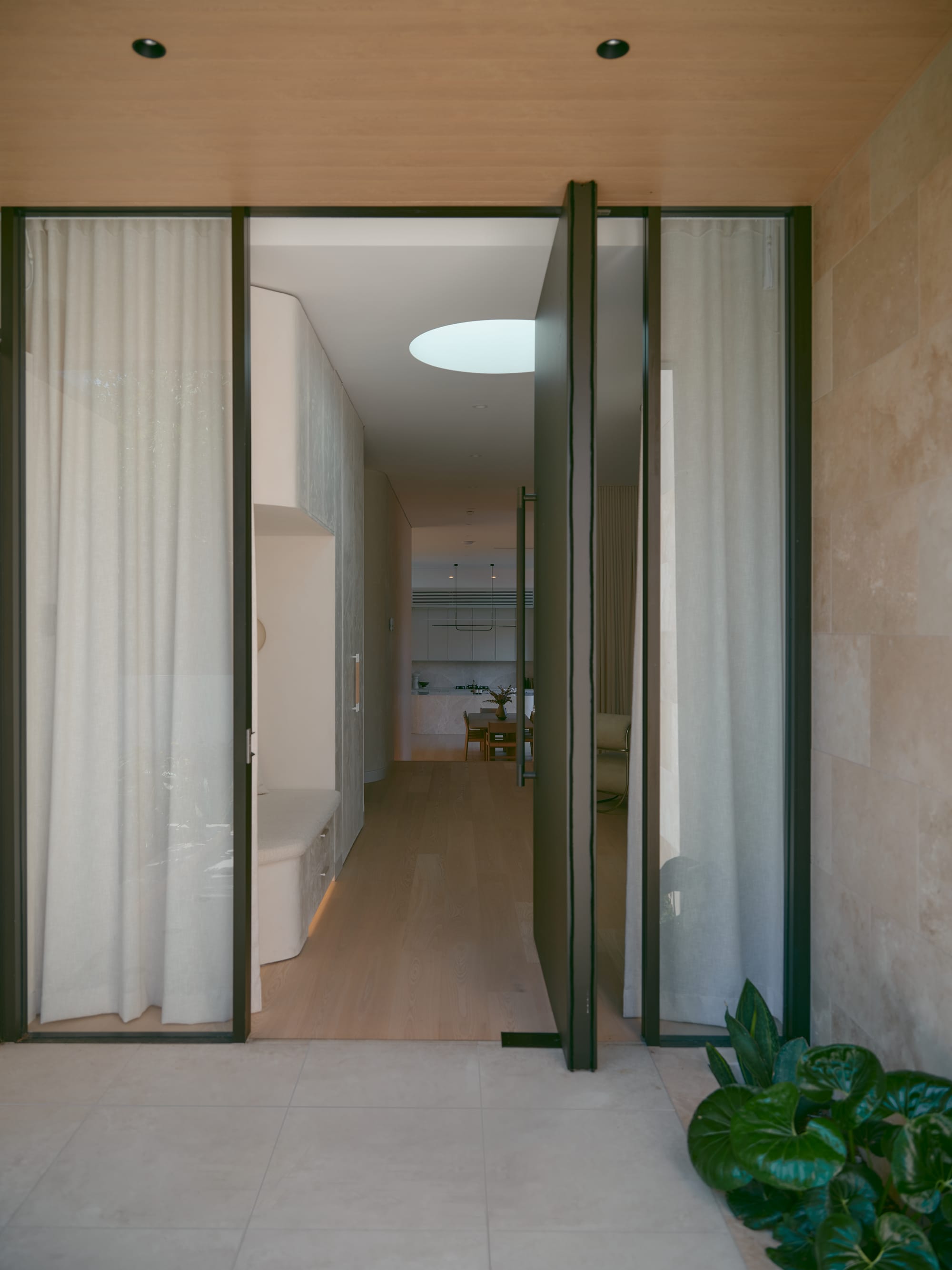 Sole House by 868 Architects. Photography by Martin Siegner. Interior view through large pivot door showing a hallway with timber flooring, soft curtains, curved joinery, and a circular skylight illuminating the entry space.