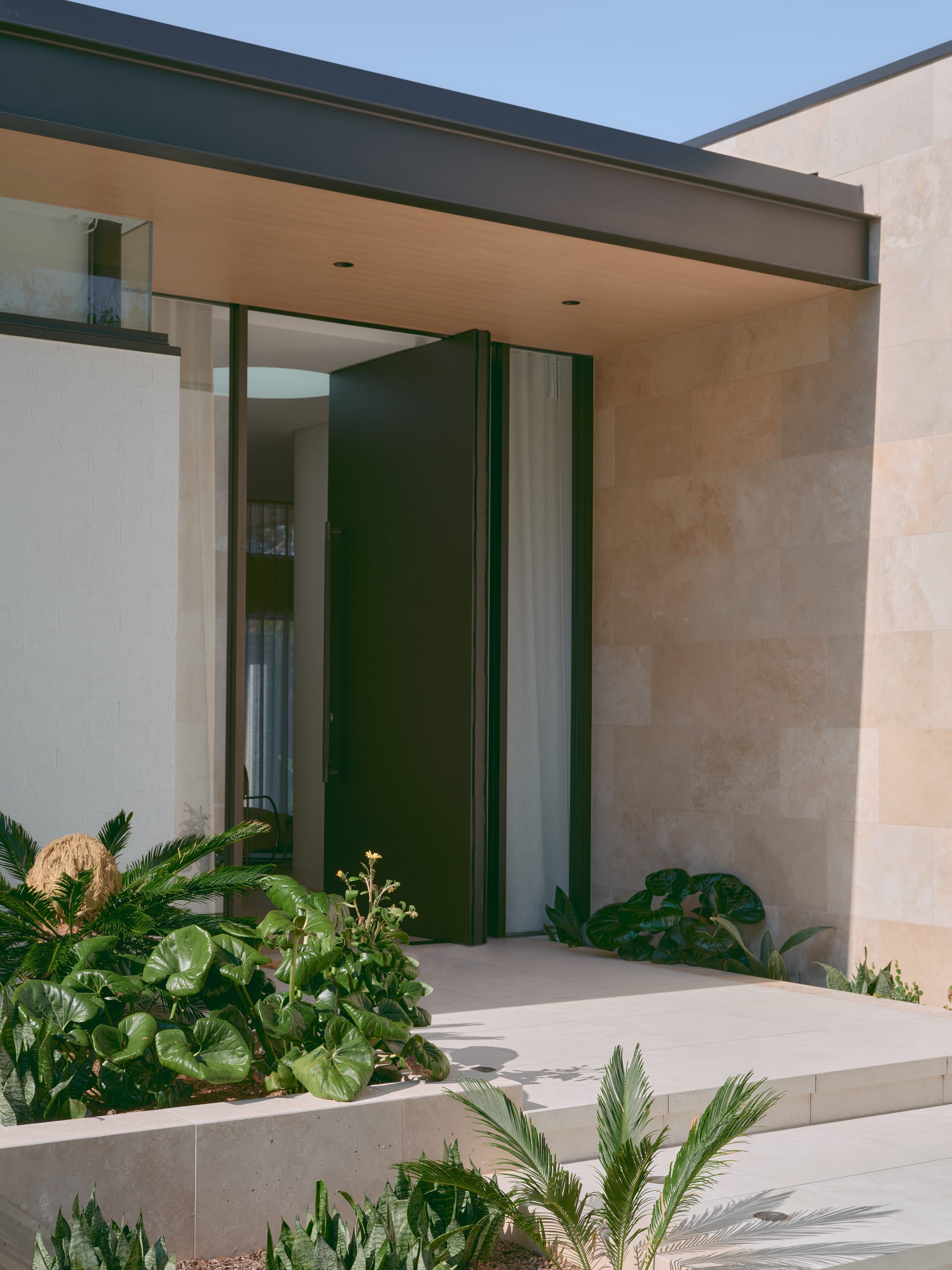 Sole House by 868 Architects. Photography by Martin Siegner. Minimalist home entrance with large pivot door, limestone cladding, and lush tropical planting.