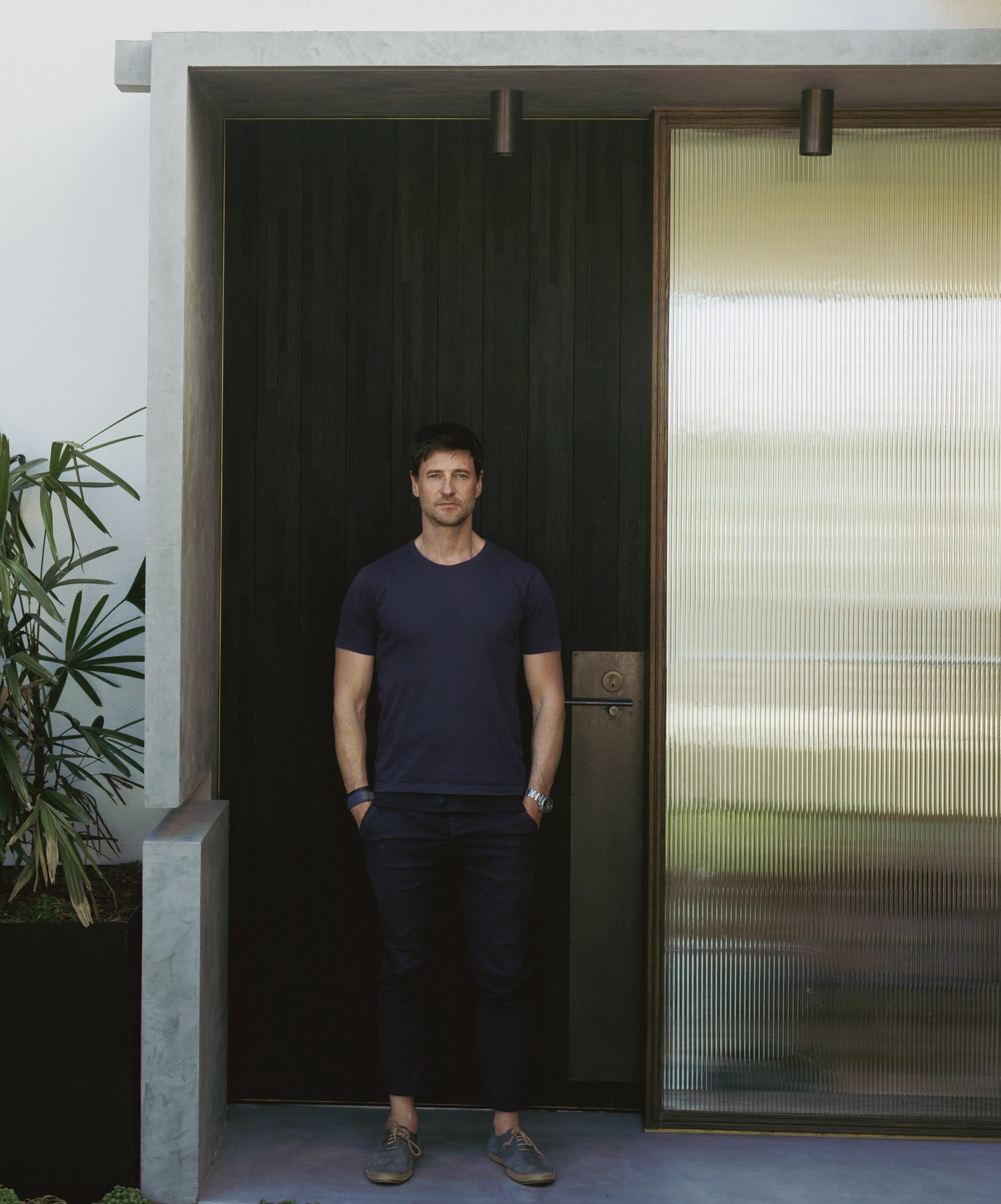 Peter Ahern by buck&simple. Photography by Tim Pascoe. Man with black hair wearing a navy shirt and black trousers standing in front of black timber door.