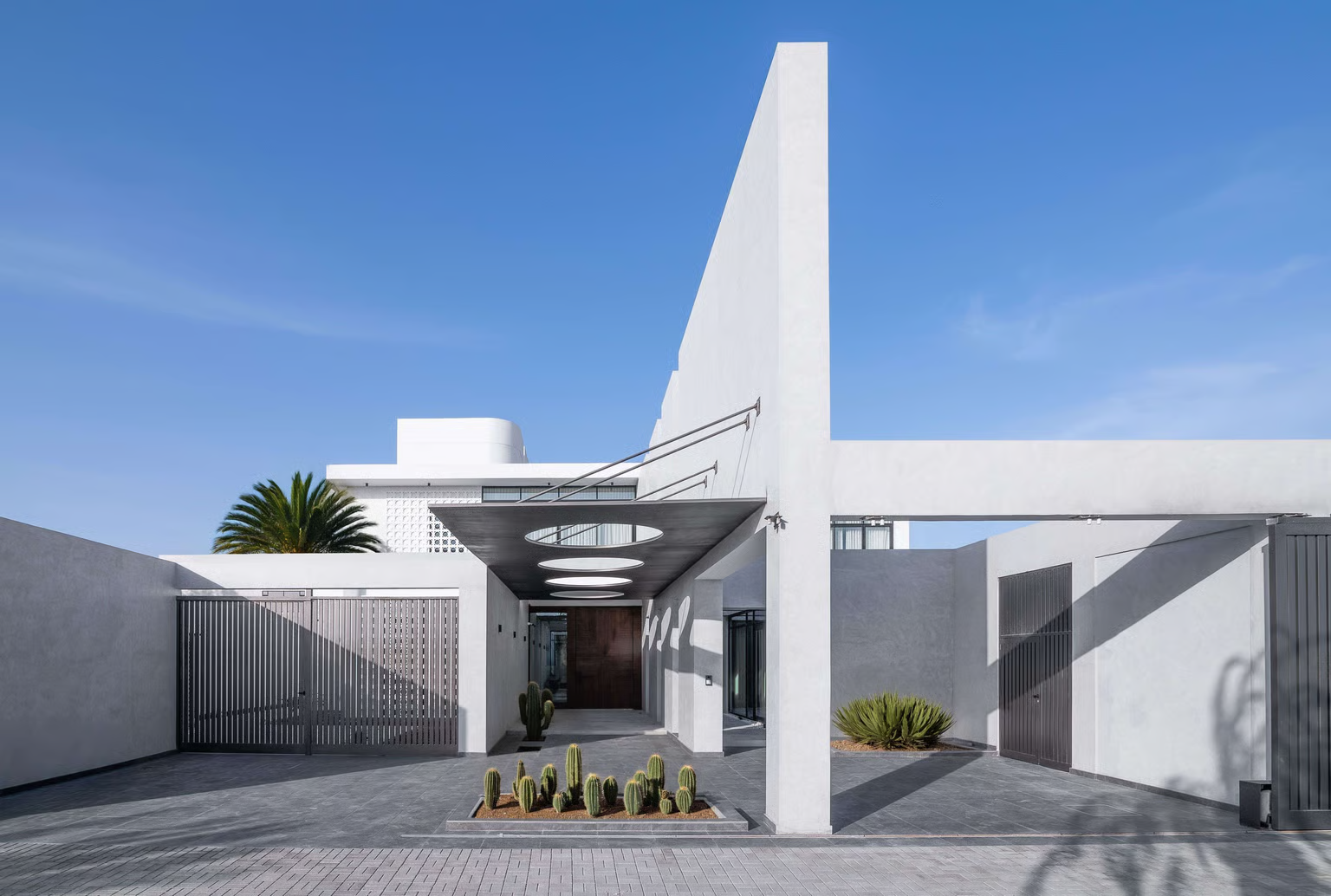 Veil House By Enclave Architects. Photography by Hassen Arante. Modern minimalist house facade featuring geometric forms, white plaster walls, a dramatic vertical fin wall, and a central entrance canopy with circular cutouts casting shadows over a cactus garden.