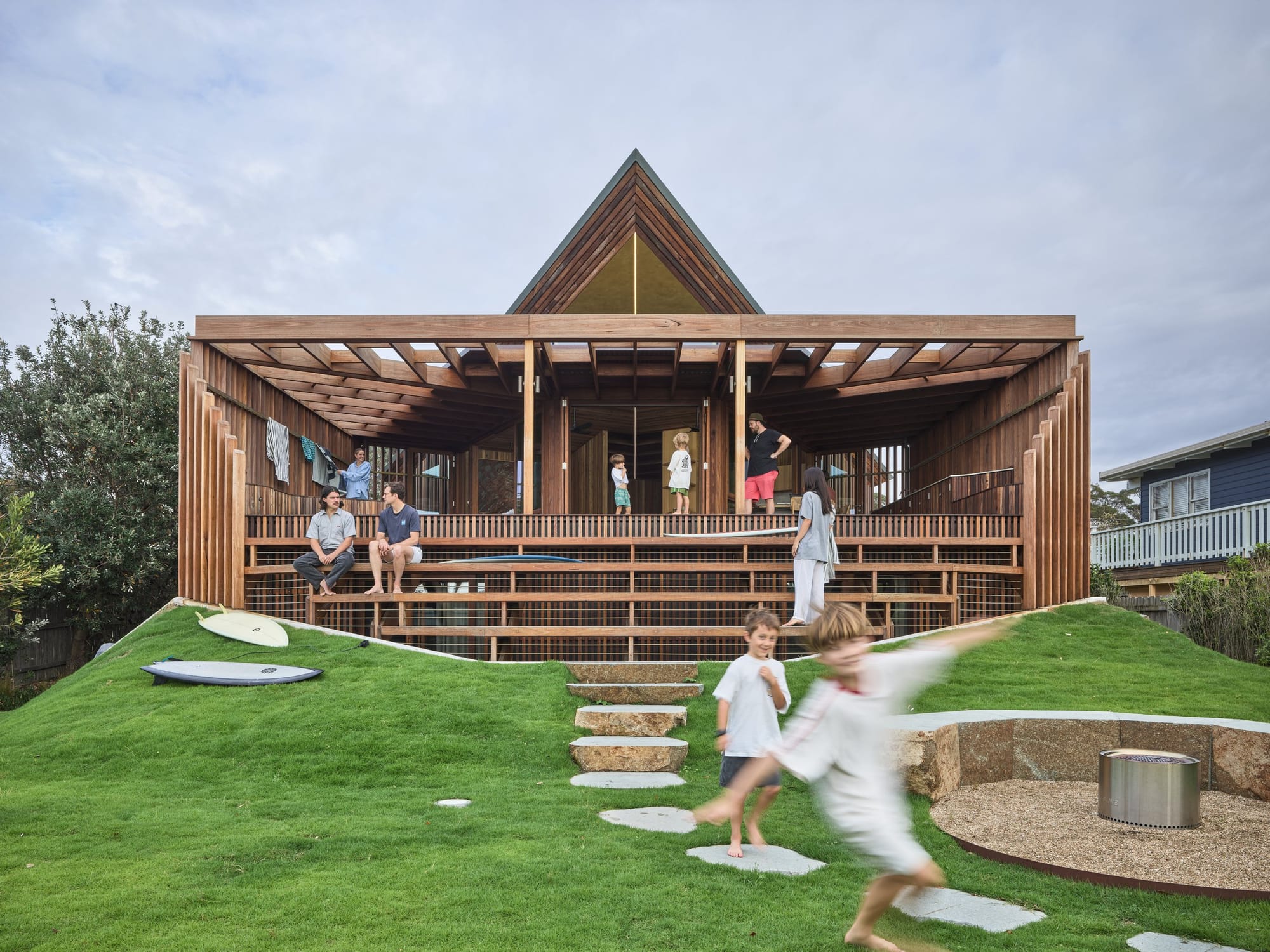 Cake House by Alexander Symes Architect. Photography by Barton Taylor. Children playing at front yard of wooden structure house with inclined landscape.