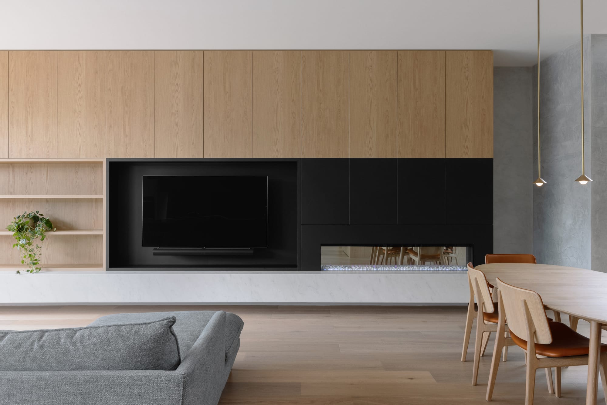 Auburn Residence by Chan Architecture. Photography by Elise Scott. Open plan living and dining space with timber cabinetry and integrated fireplace stretching the length of the back wall, with timber dining table and a grey couch in front. 