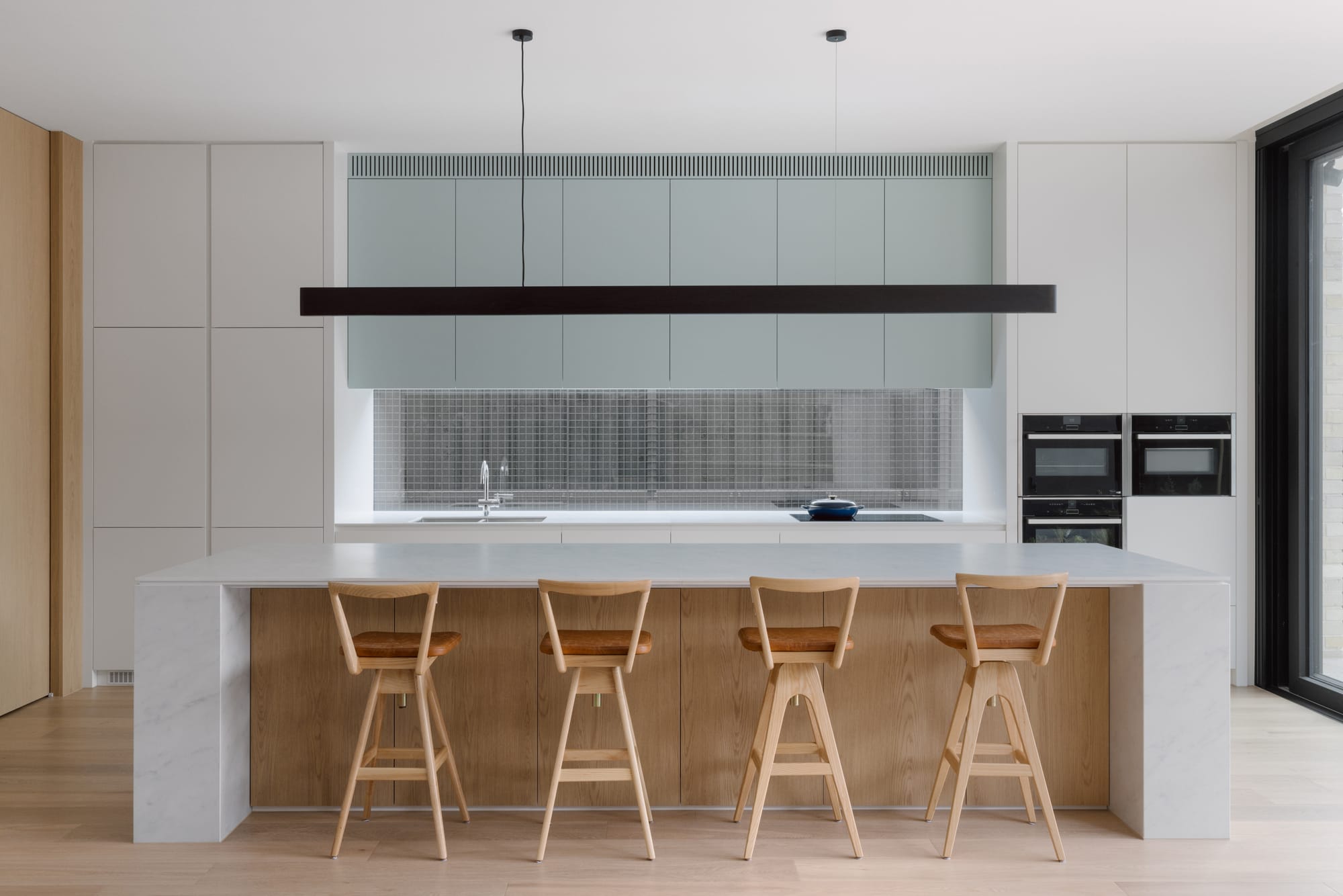 Auburn Residence by Chan Architecture. Photography by Elise Scott. Island bench with white marble benchtop, timber kickboard and timber stools in open plan kitchen with blue and white cabinetry. 
