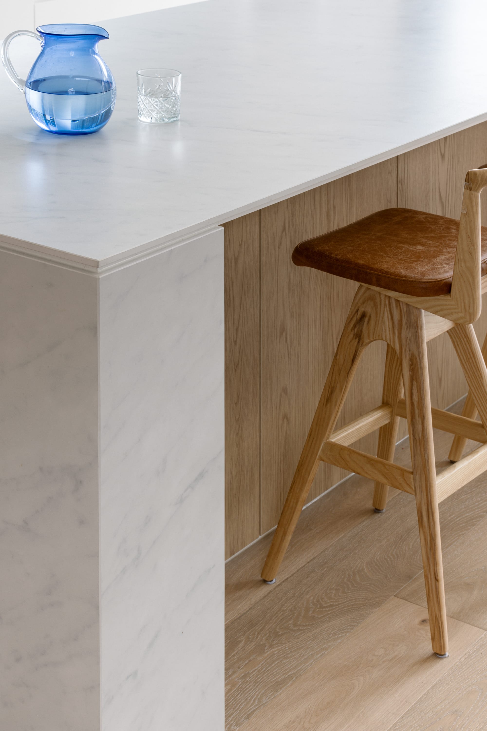 Auburn Residence by Chan Architecture. Photography by Elise Scott. Close shot of corner of white marble benchtop in kitchen with a blue water jug, contrasted against the pale timber kickboard, flooring and stool. 