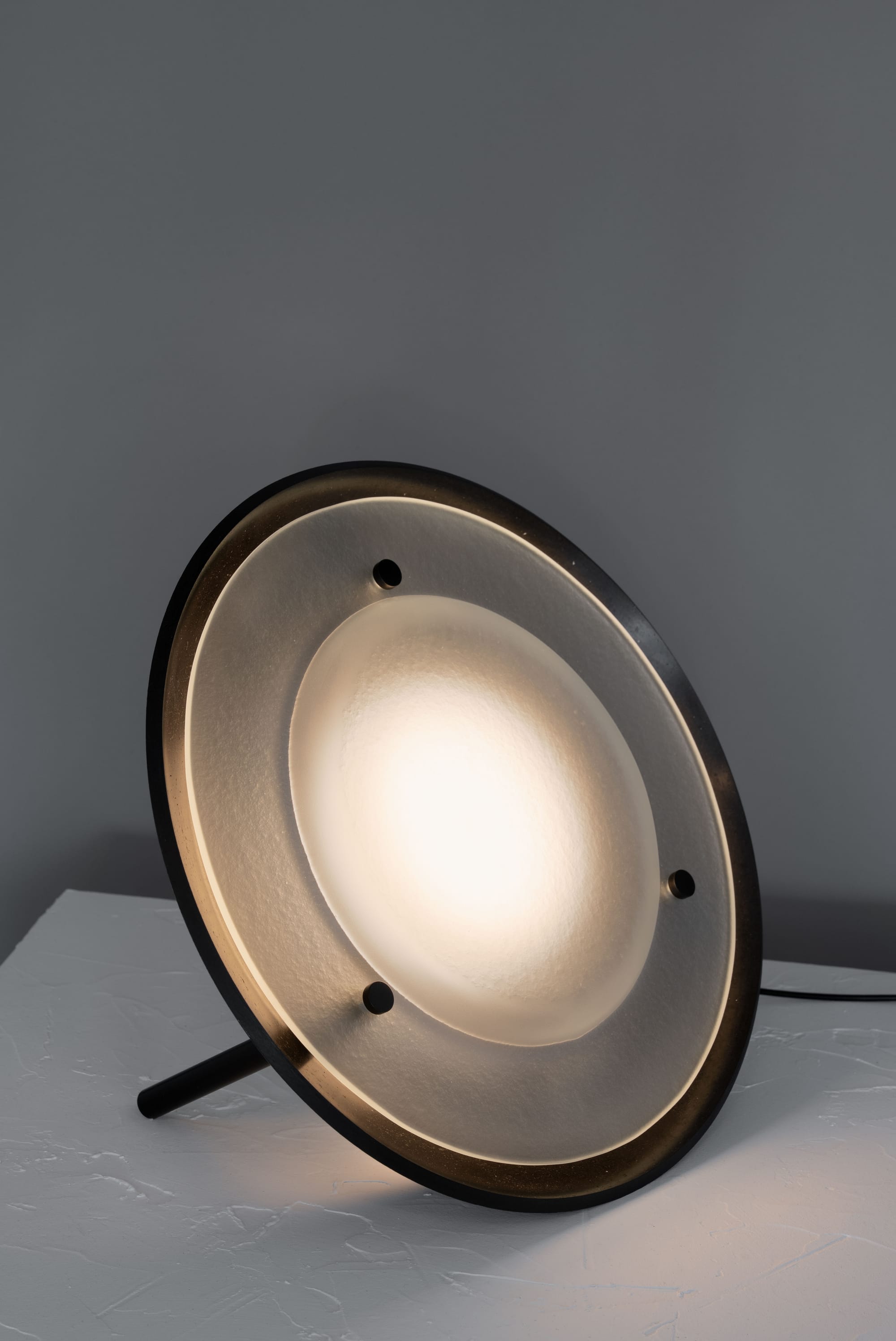 Husk Collection By South Drawn. Photography by Reannon Smith. A soft-glowing disc light rests gently on a tripod support, showcasing the Husk Collection’s tactile finish and sculptural simplicity.