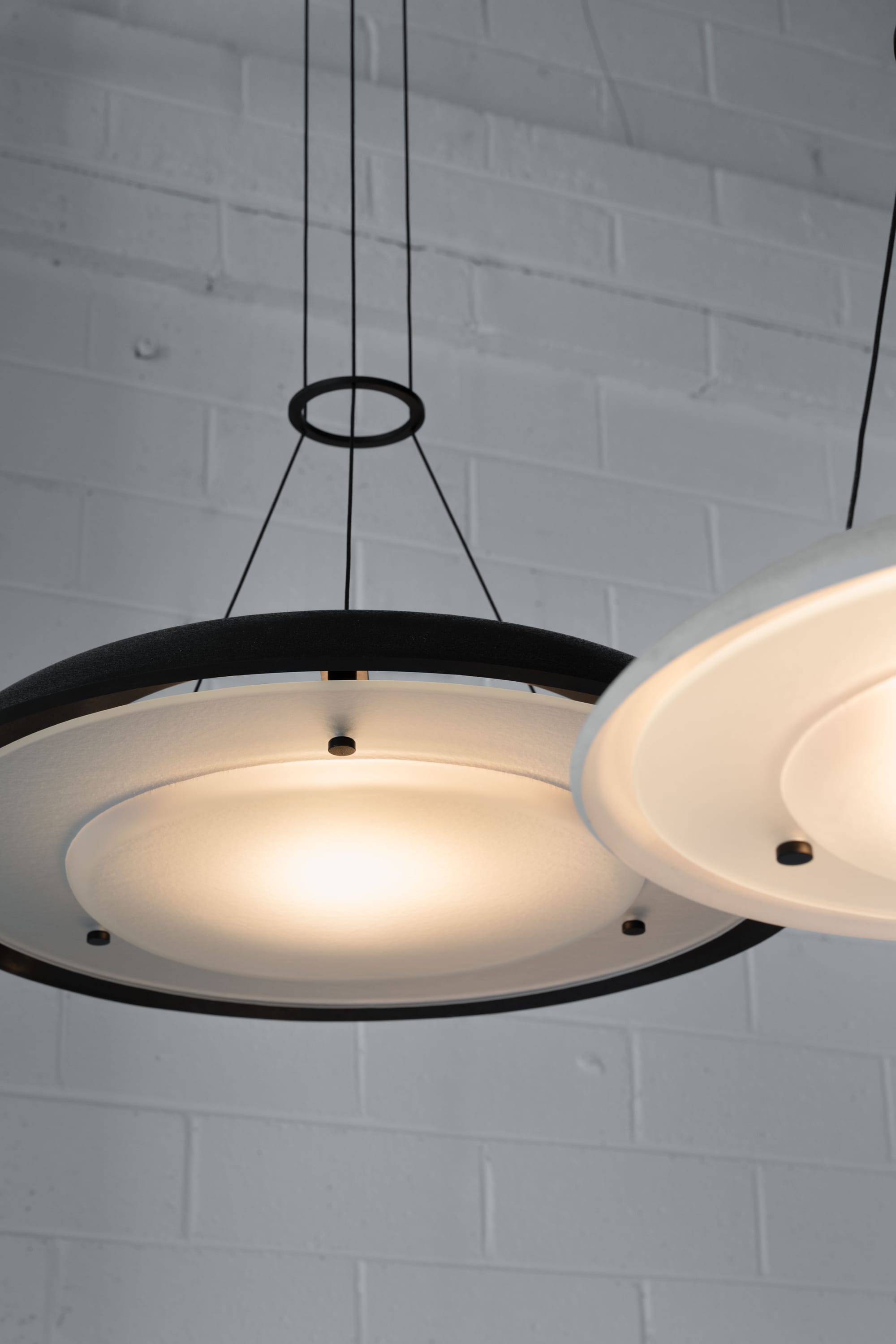 Husk Collection By South Drawn. Photography by Reannon Smith. Two suspended disc-like luminaires glow softly against a matte white brick wall, highlighting their refined detailing and layered construction.