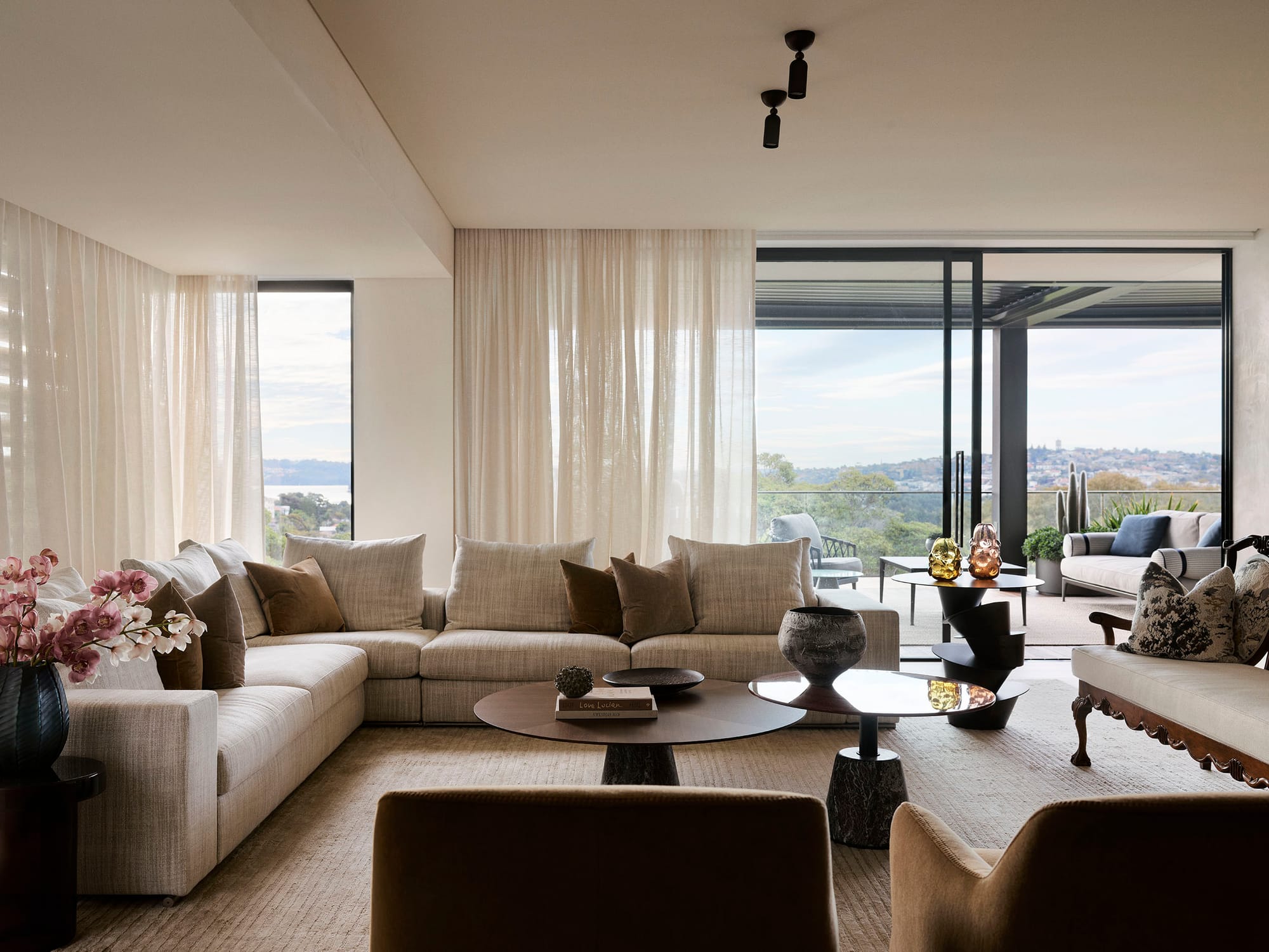 Bundarra Penthouse by Marylou Sobel Interior Design. Photography by Anson Smart. Elegant living room with a neutral-toned sectional sofa, layered coffee tables, sheer curtains, and expansive glass doors opening to a terrace with panoramic views.