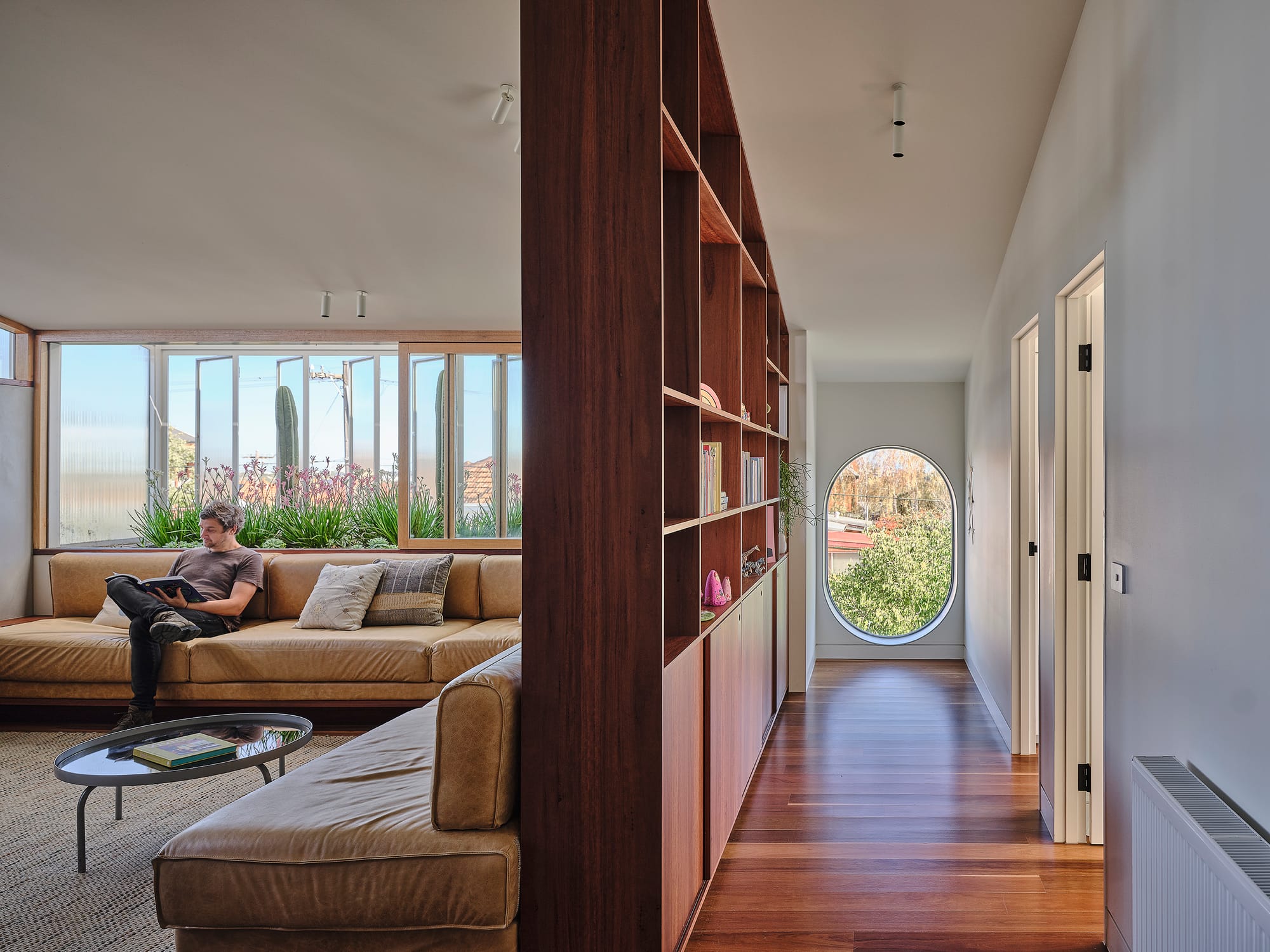 Salmon House by True Story. Photography by Dean Bradley.Split space showing a living area with tan built-in sofas and large corner windows with cacti, and a hallway with timber shelving and a round window at the end.