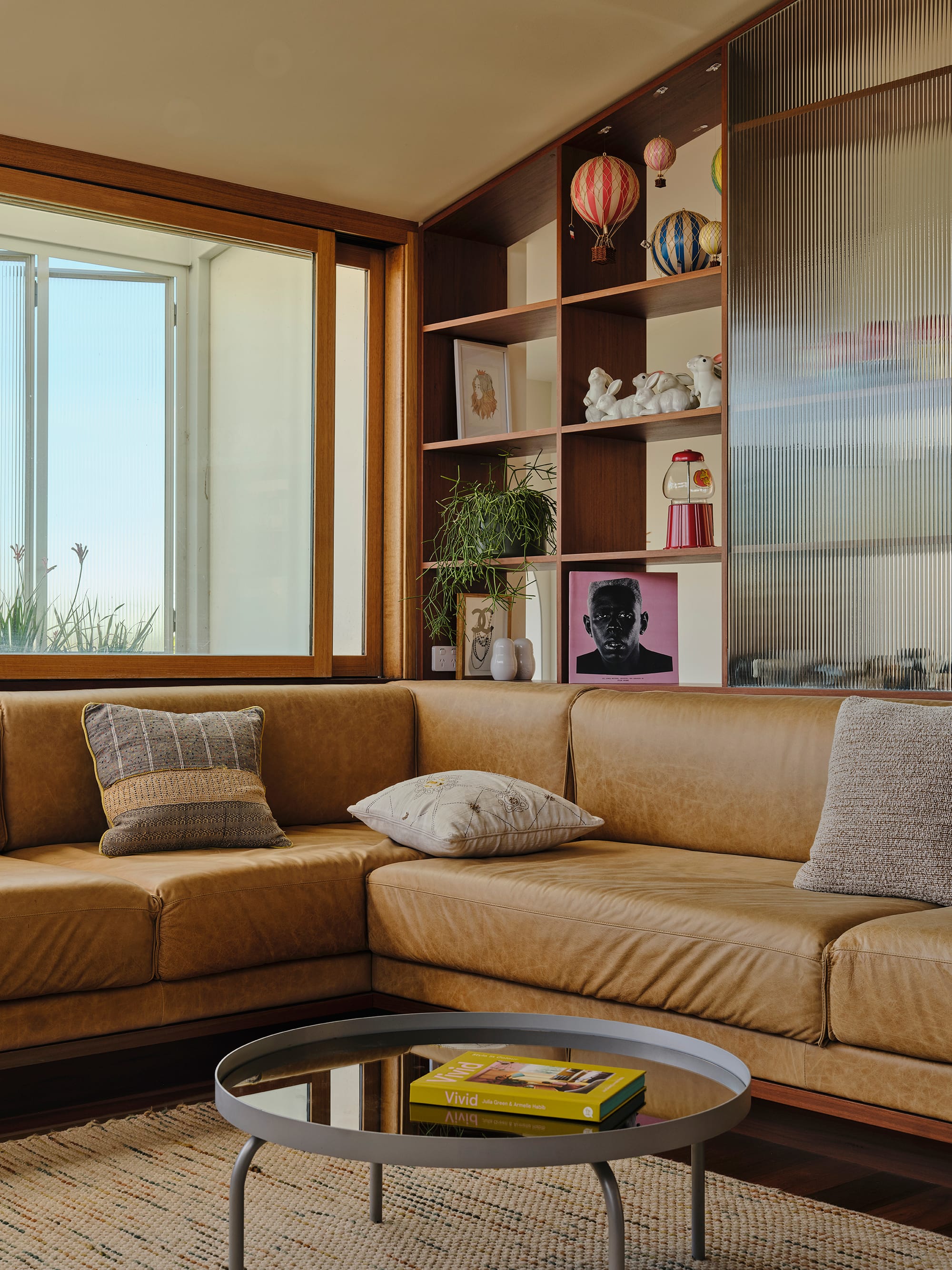 Salmon House by True Story. Photography by Dean Bradley.Close-up of a cozy tan leather sectional in a living room with a display shelf featuring playful figurines, books, and art, set beside a large window and fluted glass screen.