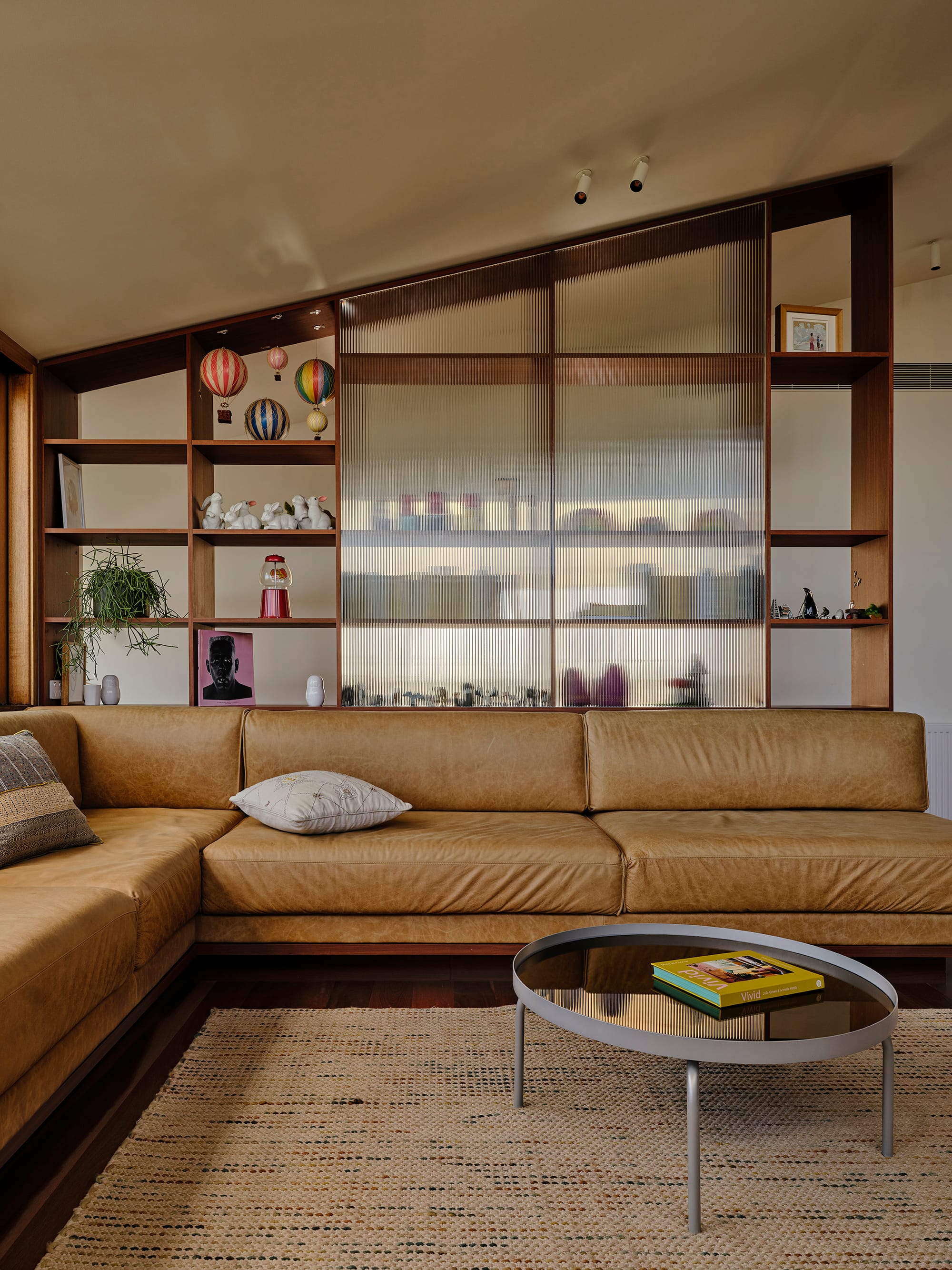 Salmon House by True Story. Photography by Dean Bradley.Caramel leather sofa in front of timber shelves with playful objects and fluted glass panels.