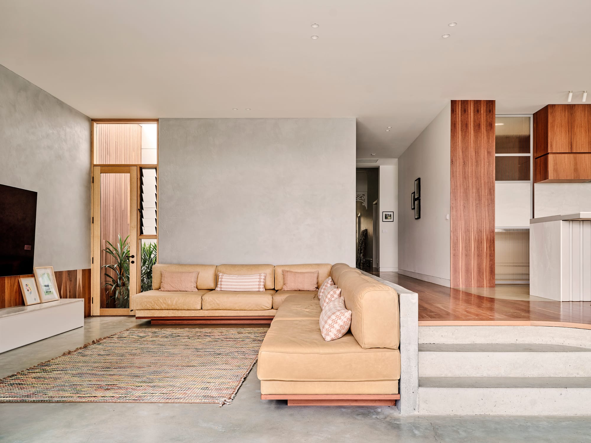 Salmon House by True Story. Photography by Dean Bradley.A sunken living room with a beige sectional sofa, textured rug, and timber finishes connects to an elevated kitchen with warm wood cabinetry.